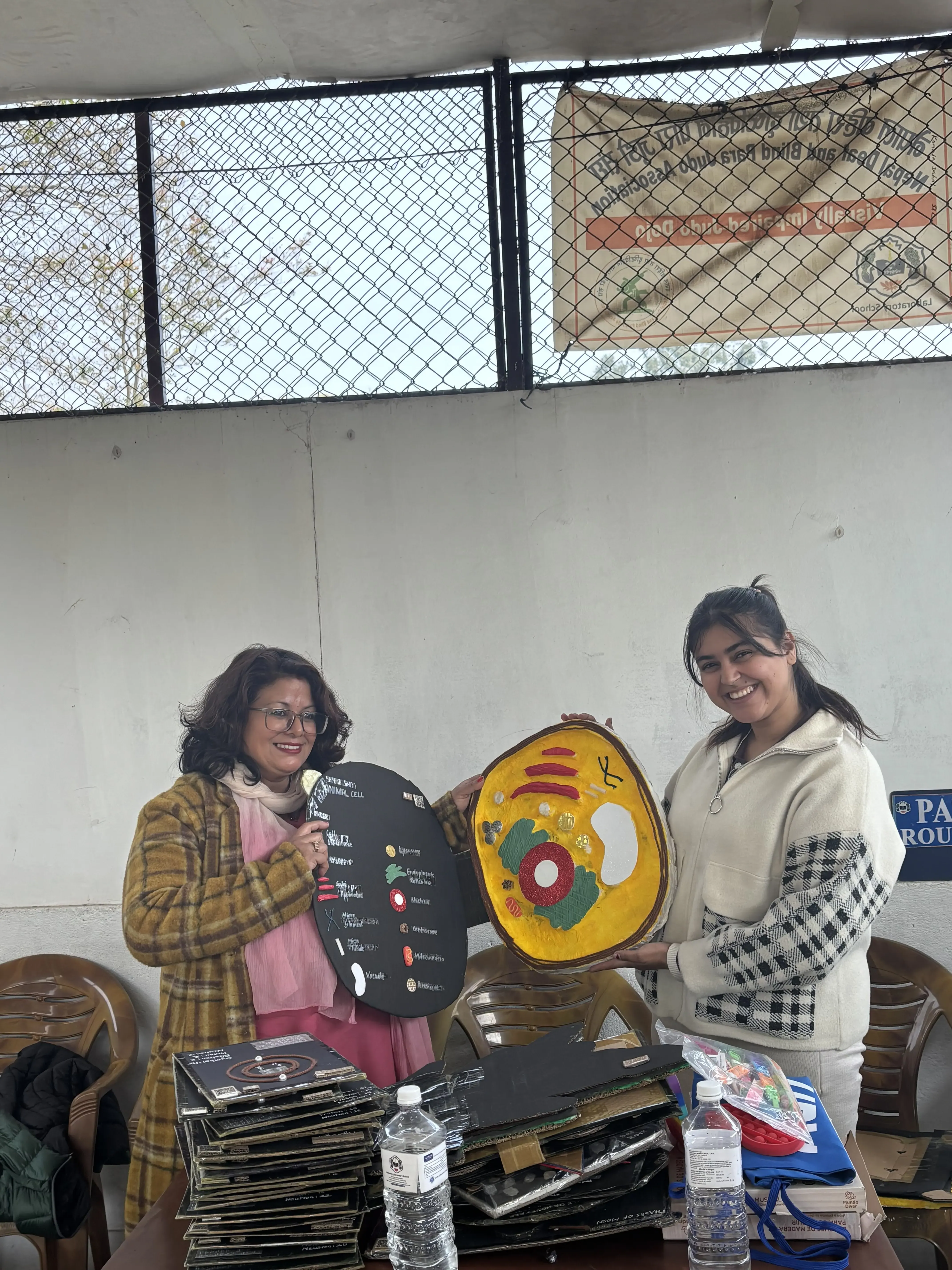 Two women are standing indoors near a table, smiling and holding up large tactile 3D educational models. The woman on the left is displaying a 3D model of plant cell. The woman on the right is holding a 3D model of animal cell. On the table in front of them, several more stacked models, water bottles, and other materials are visible. They are participating in a formal event to hand over accessible 3D models to the school.