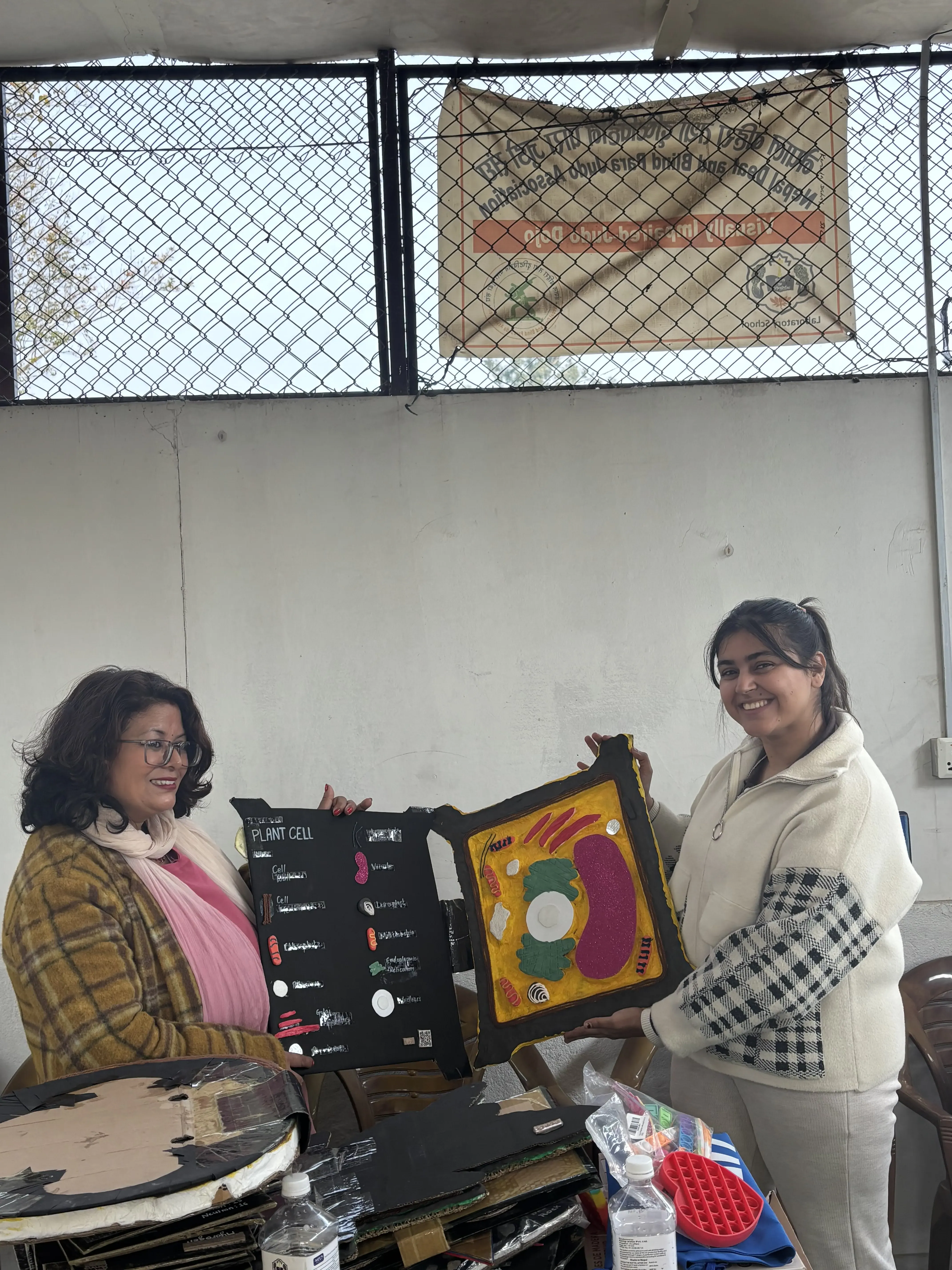 Two women are indoors and smiling as they display a large, tactile 3D educational model of a plant cell. The woman on the right, dressed in a light-colored outfit, is holding the colorful model, which shows the cell's internal structures in raised sections for touch-based learning. The woman on the left, wearing a yellow-checkered coat and scarf, is holding a corresponding black model with labeled parts, including braille text for accessibility. The table in front of them has more educational models and supplies.