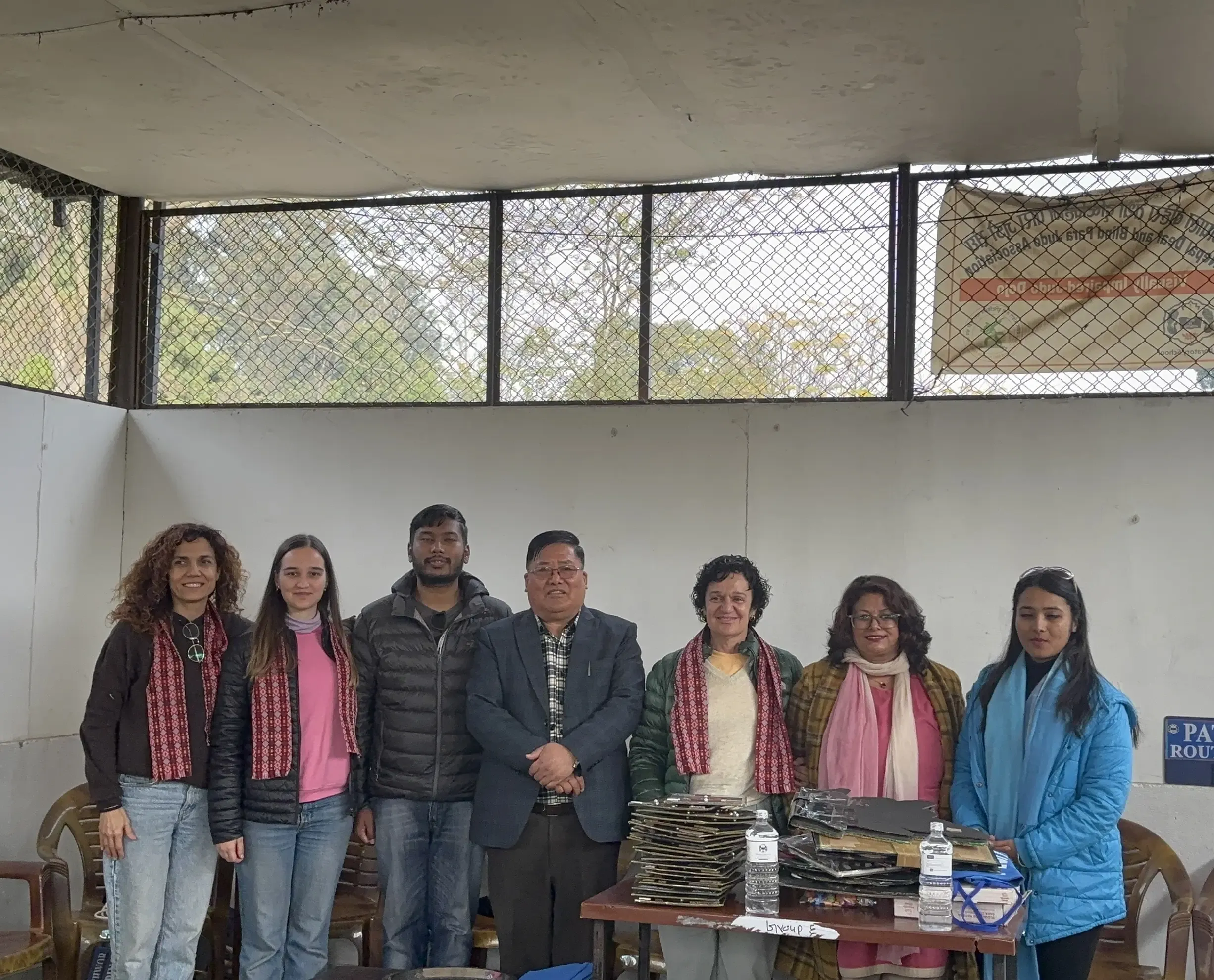 A group of seven adults—four women and three men—are standing together indoors, posing for a group photo behind a table. Several of the women are wearing patterned scarves, and some of the group members are dressed in warm jackets and coats. On the table in front of them, there are stacks of tactile 3D educational models and a few bottles of water, suggesting a recent event involving educational materials. The group stands in front of a large window with a mesh screen, letting in natural light. The atmosphere is formal and friendly, marking the conclusion of a handover event.
