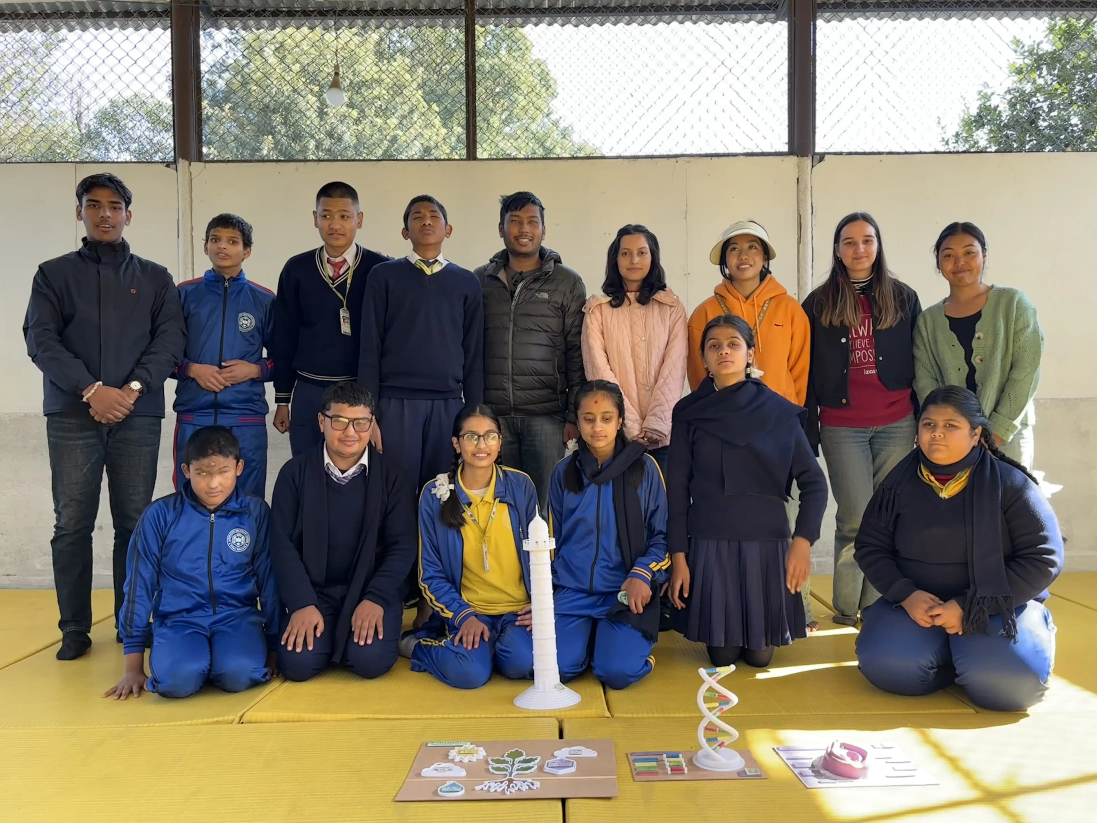 Group photo of students and adults taken indoors on yellow mats, in front of a wall with wire mesh windows that let in natural light with trees visible outside. The group includes children in blue and navy school uniforms and adults in casual winter clothing. Some students and teachers are standing in the back row while others are sitting or kneeling in the front. In front of the group, there are educational models on display: the Dharahara tower, a DNA double helix, female external reproductive organ, and photosynthesis.