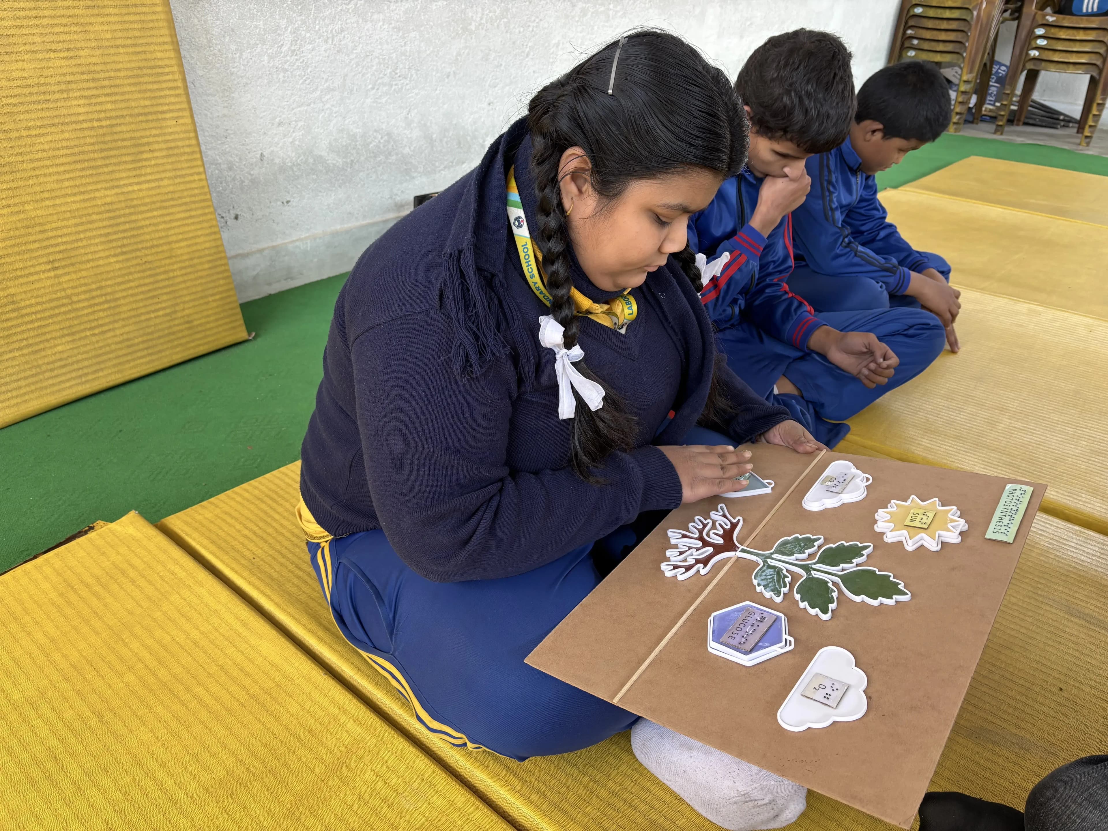 A girl in a school uniform (navy sweater, blue pants with yellow stripes, and a scarf) is sitting on a yellow mat, engaged with a tactile educational board. The board has raised, colorful 3D pieces depicting a diagram of a plant with labeled parts such as leaves, sun, and possibly water drops, likely a photosynthesis educational model. Two other students in similar uniforms are sitting next to her, also on the yellow mats, observing and waiting their turn.