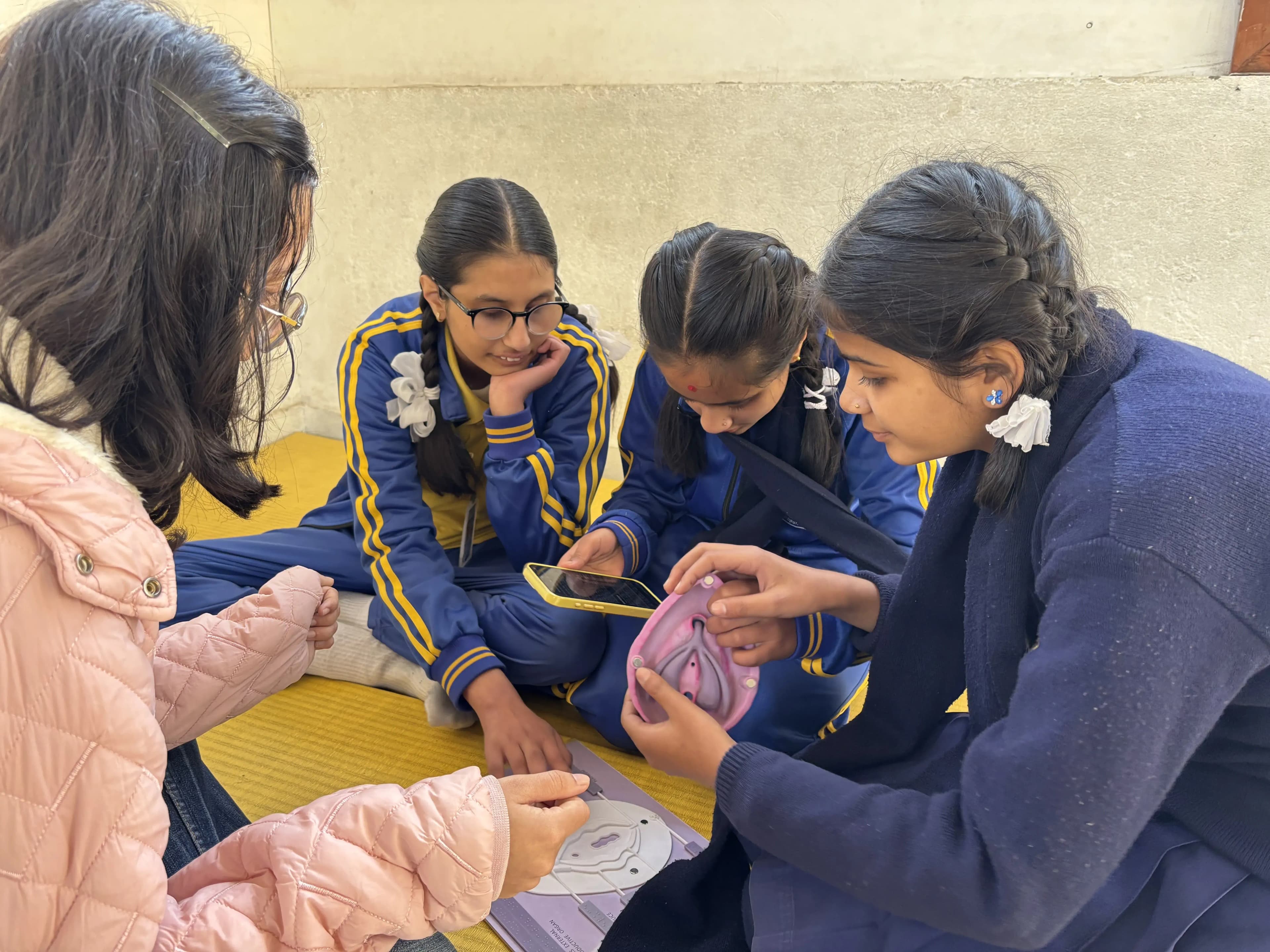 Four girls sitting on a yellow mat against a light-colored wall, engaged in a hands-on educational activity. Two of the girls are wearing blue tracksuits with yellow stripes, and one is wearing a navy school sweater and skirt. One girl in the navy sweater is holding a detailed anatomical model of human female reproductive organs, while another girl beside her is holding a phone, using it for its audio description from CFC app. A paper or board featuring a labeled diagram rests on the mat in front of them. The fourth girl, wearing a light pink quilted jacket and glasses, is observing and participating in the discussion.