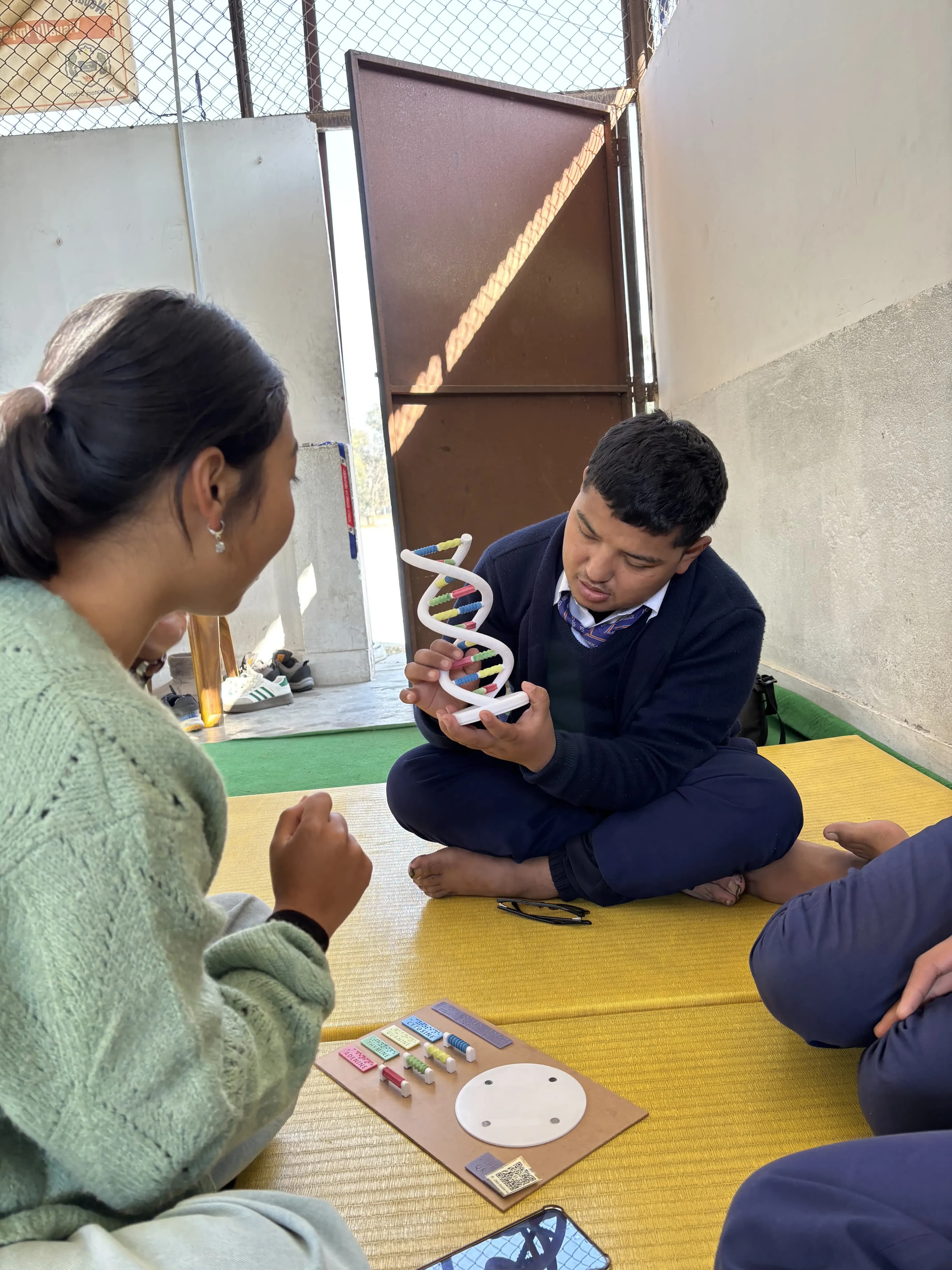 A boy in a navy school uniform is sitting cross-legged on a yellow mat near a wall, holding and closely examining a 3D model of a DNA double helix. In the foreground, a girl in a light green sweater is also seated on the mat, watching him and interacting with a tactile educational model laid out between them. Another person, only partially visible, is sitting nearby.