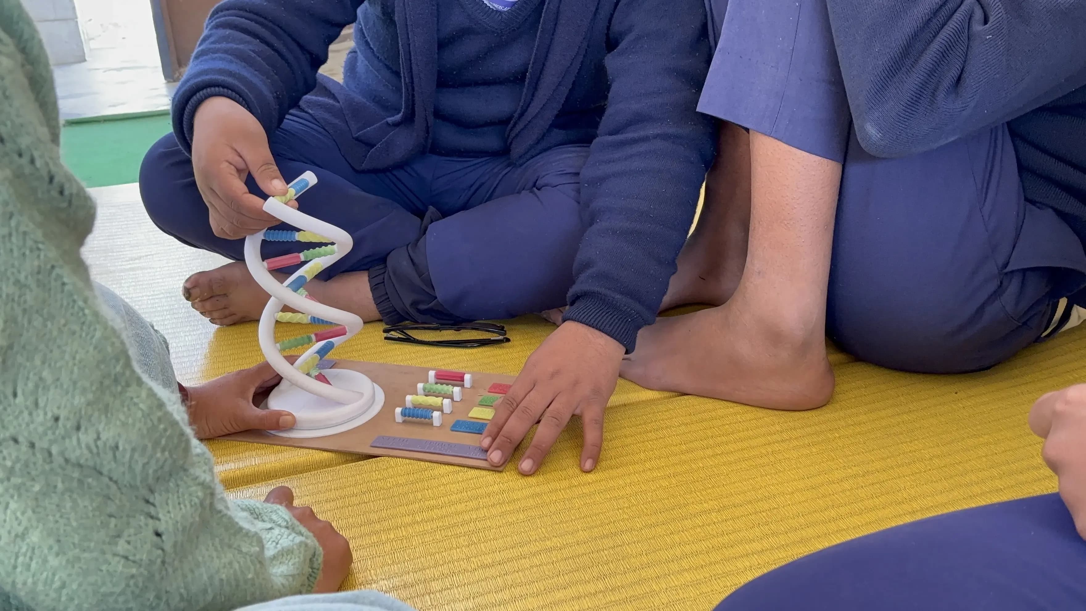 Children sitting on a yellow mat, closely interacting with a 3D educational model of a DNA double helix and a molecular learning kit. The children are wearing blue clothing and seem to be engaged in a workshop.