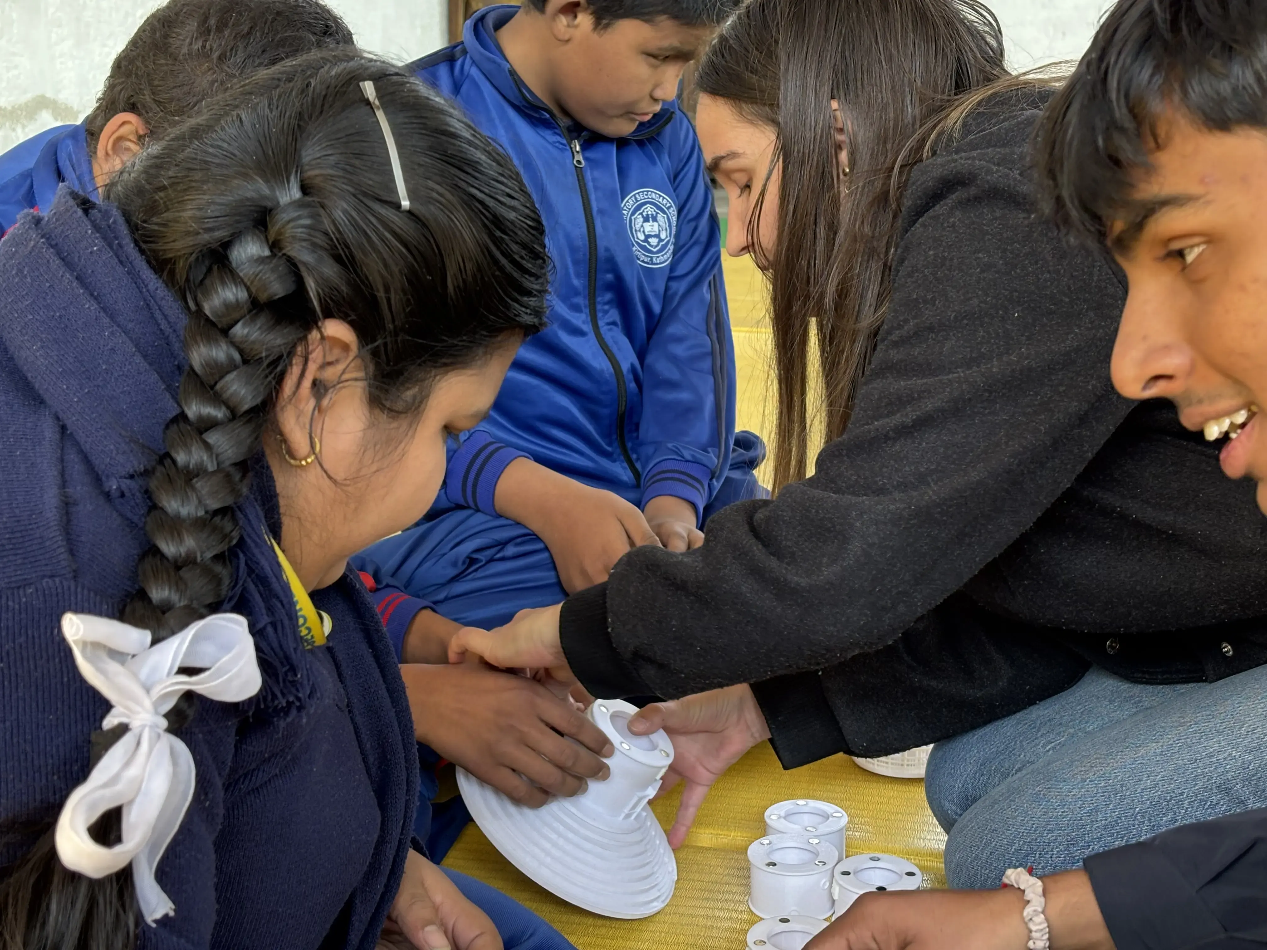 Group of students and a project member closely gathered on a yellow mat, working hands-on with model of Dharahara Tower. The participants, wearing blue uniforms and winter clothes, appear engaged and focused on assembling or exploring the models, as part of the workshop. One girl with a braid and white ribbon is in the foreground, while others look on and participate intently.