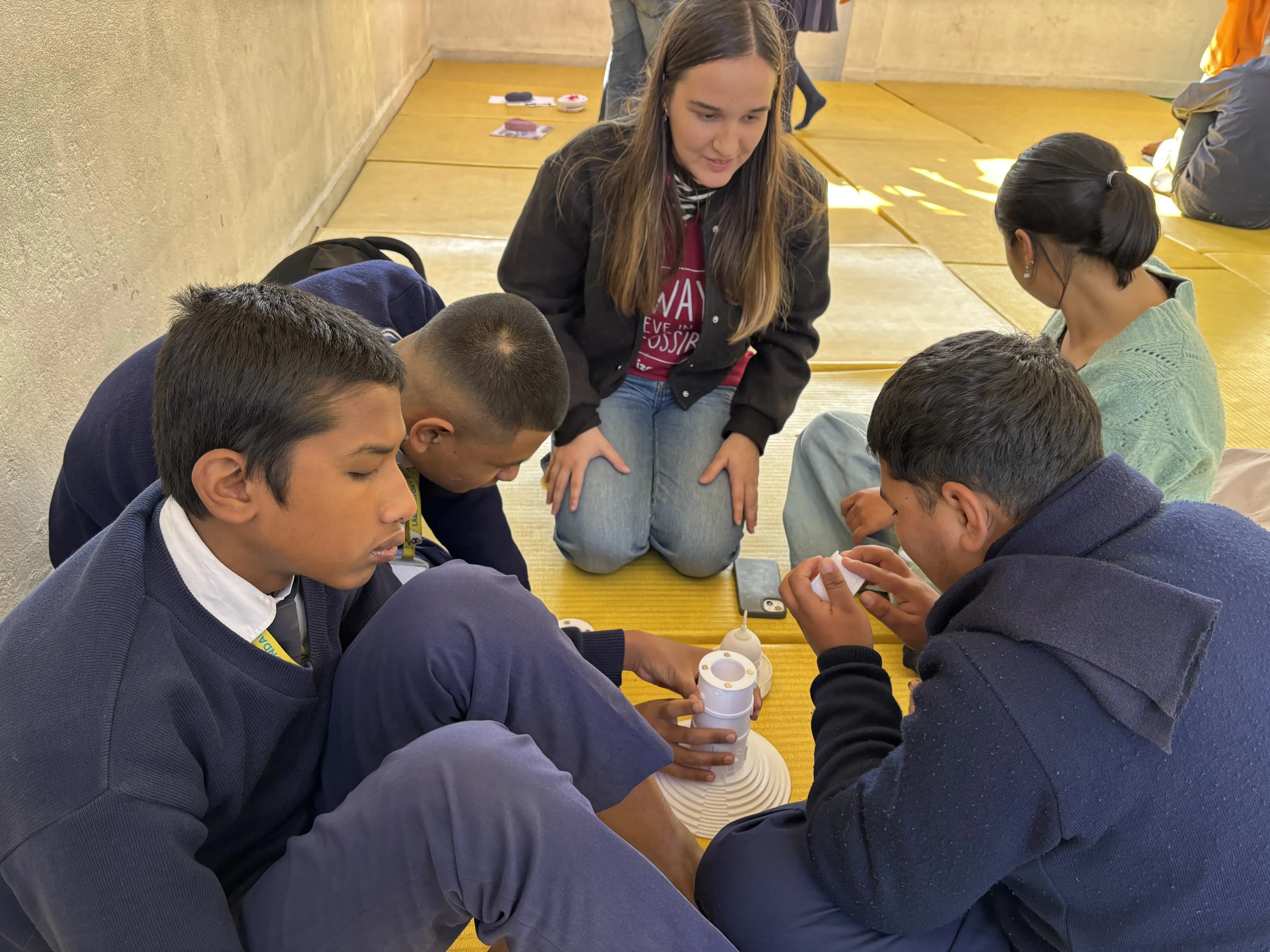 Group of students and a project member sitting in a circle on a yellow mat, actively engaged with a 3D model of Dharahara Tower that is placed on the floor. The students are in school uniforms and appear focused and curious, examining or assembling the model together. The project member, seated with them, is providing guidance and observing their interaction, creating a collaborative learning environment.