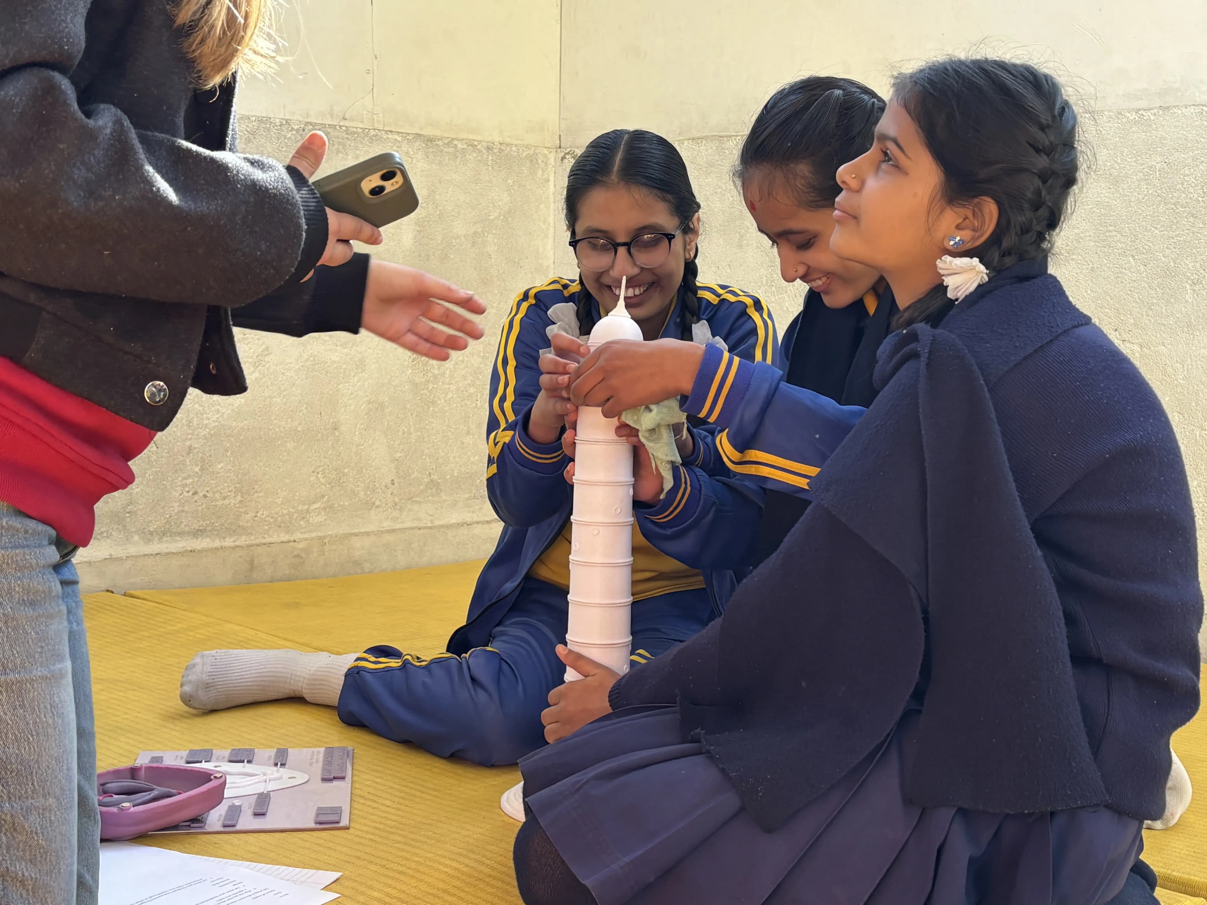 Three girls in school uniforms are sitting on a yellow mat, smiling and collaborating as they assemble a 3D model of Dharahara Tower, as part of a workshop. Another person standing nearby is holding a smartphone, possibly taking a photo or video of the activity.