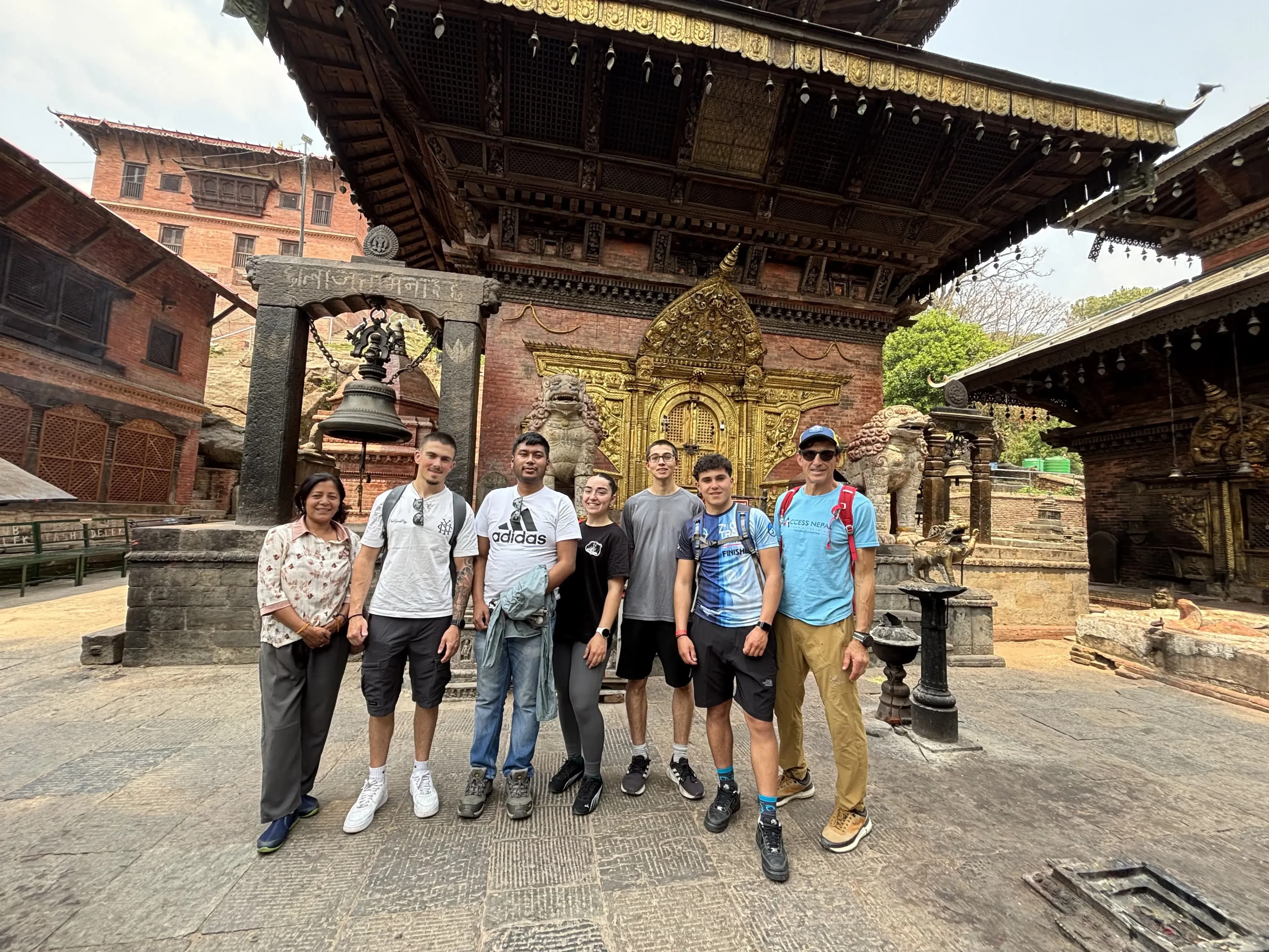 A group of seven people stand side by side posing and smiling in front of an ornate temple courtyard, with carved stone, a large hanging bell on a stone frame to the left, and an intricately decorated golden doorway and statues set into a red-brick building in the background.