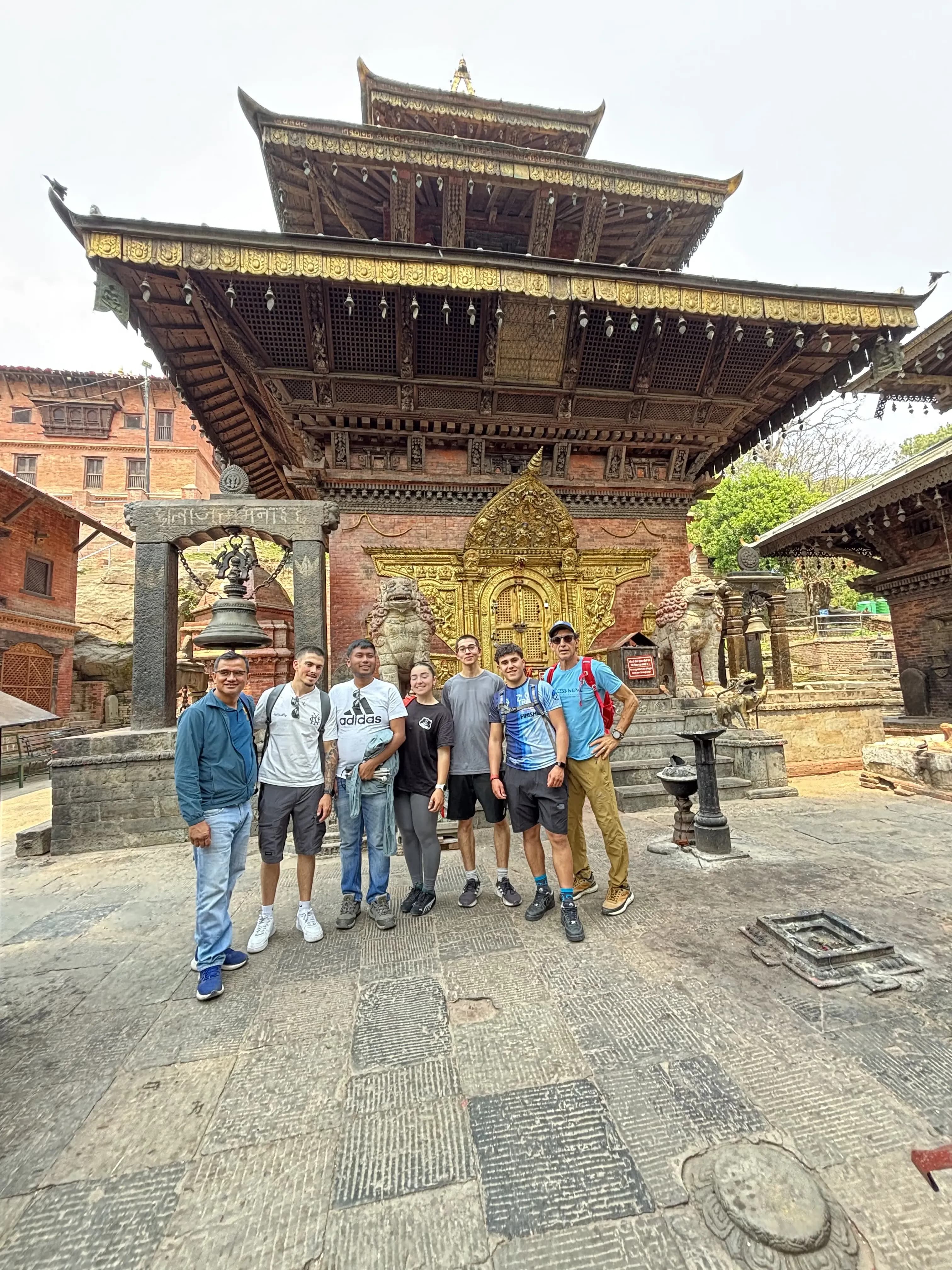 Group of eight people standing in a row and smiling in a stone courtyard, with an ornate red-brick and dark-wood temple behind them featuring a richly carved golden doorway, decorative roof carvings, and a large hanging bell on a stone frame in the foreground.