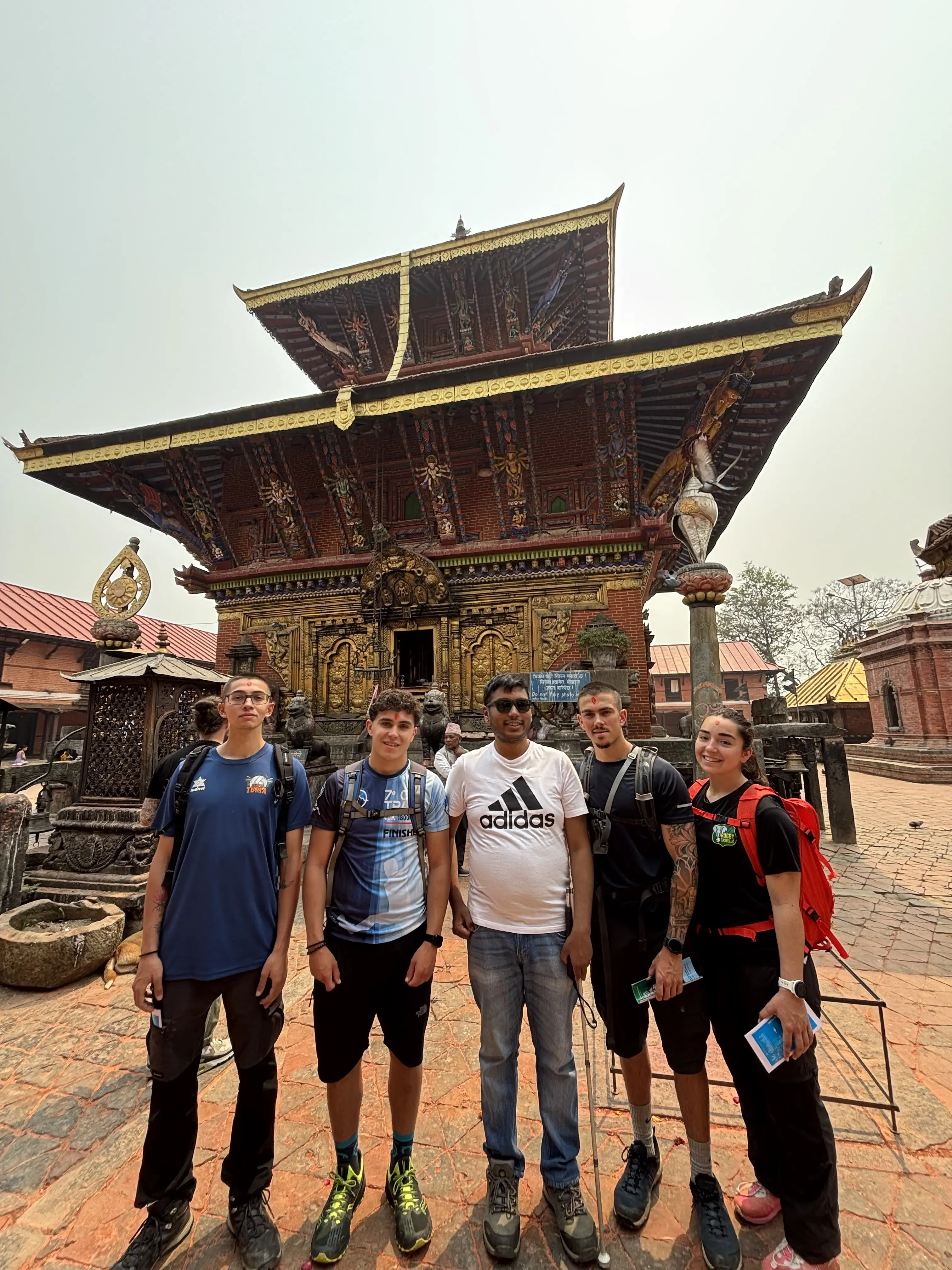 Six people stand in a loose line on a patterned stone courtyard, smiling toward the camera in front of an ornate temple with richly carved golden doors and pillars, and a tall tiered roof decorated with dark wood, red and gold details, and intricate colorful patterns.