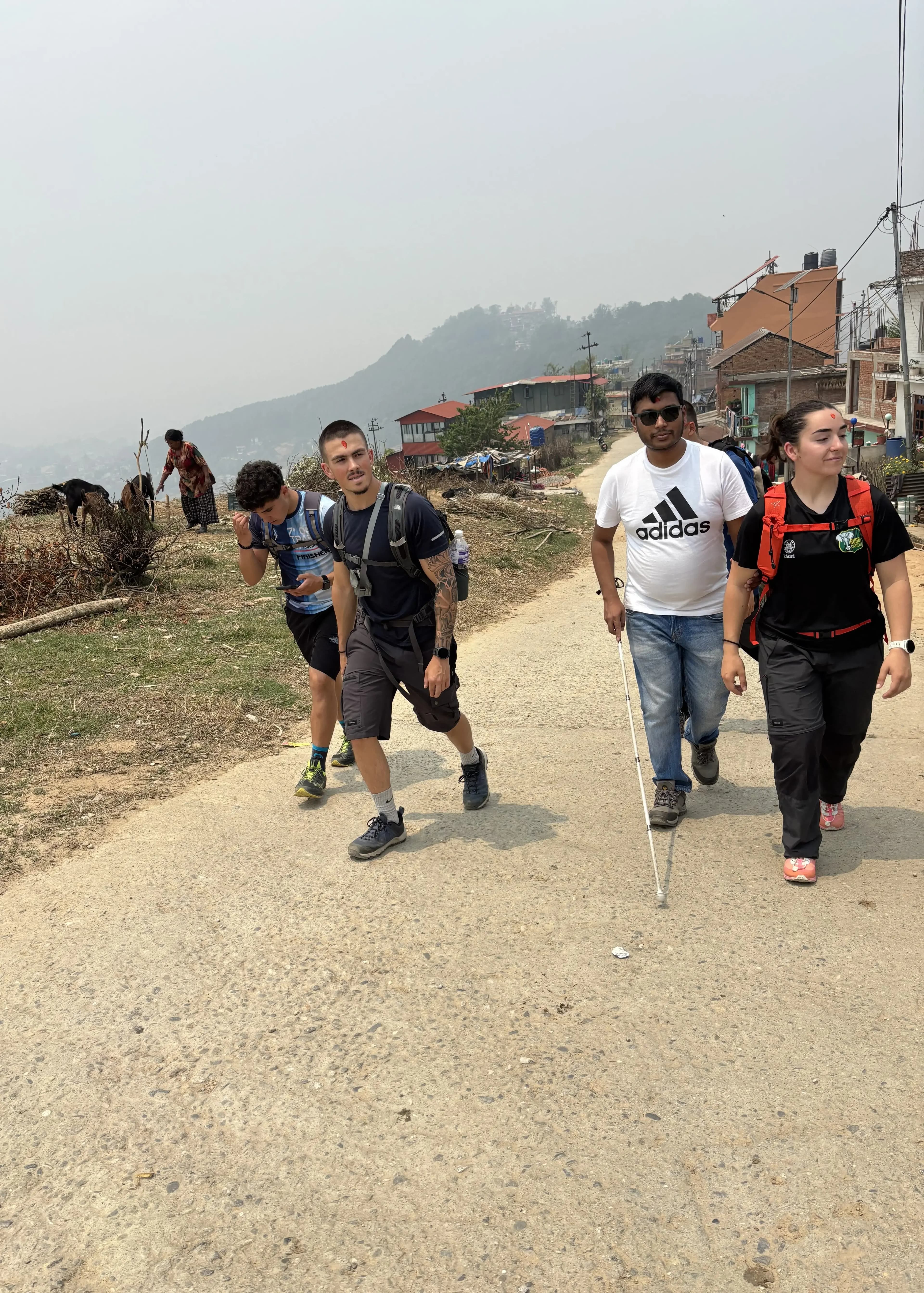 Three people with backpacks walk uphill along a wide dirt road in a rural, hilly area, smiling and talking, with simple houses and hazy green hills stretching into the distance under a bright, slightly foggy sky.
