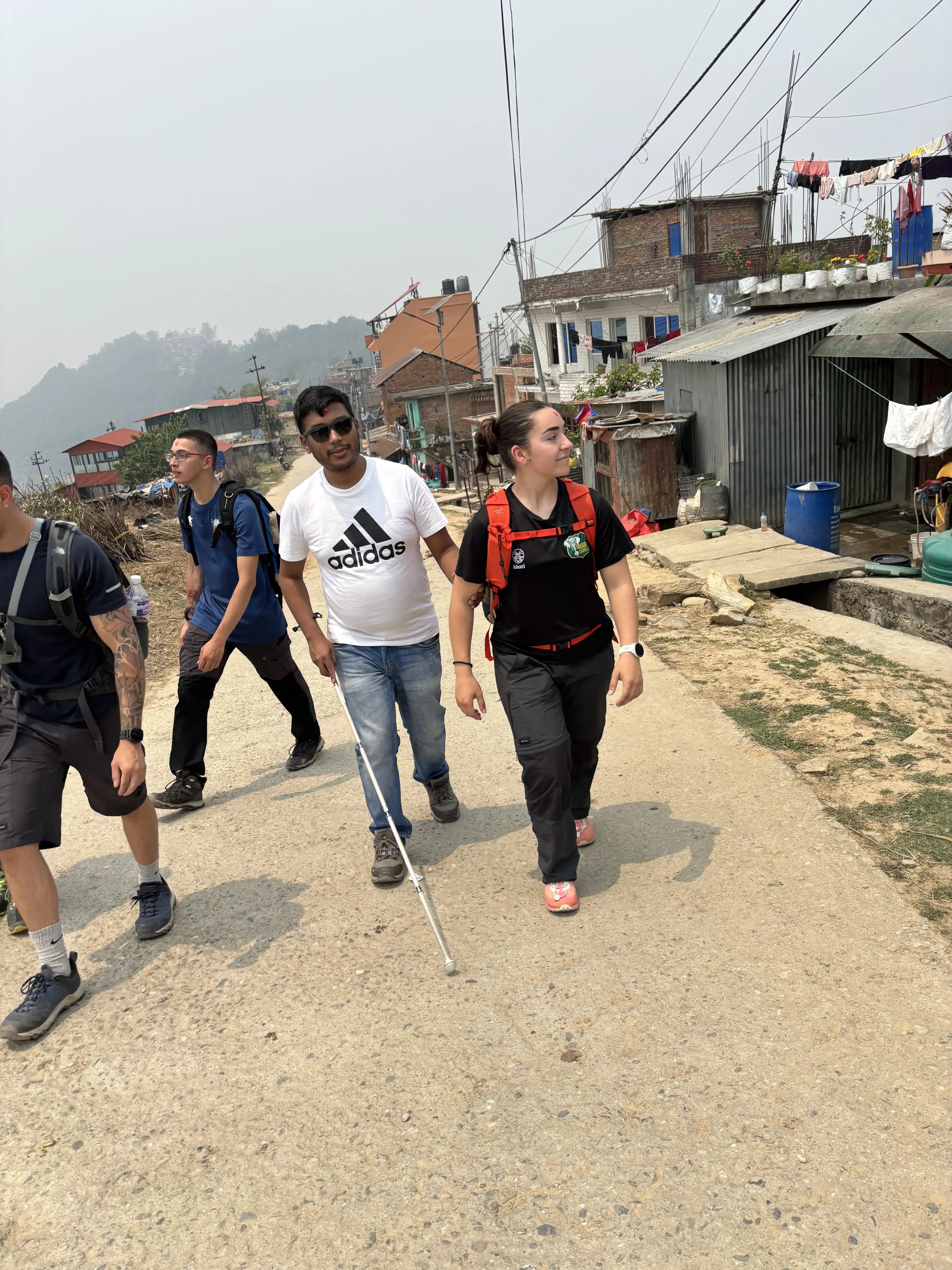 Four hikers walking together up a dusty village road, one using a white cane, all wearing casual clothes and backpacks, with simple houses, utility wires, and hazy green hills in the background.