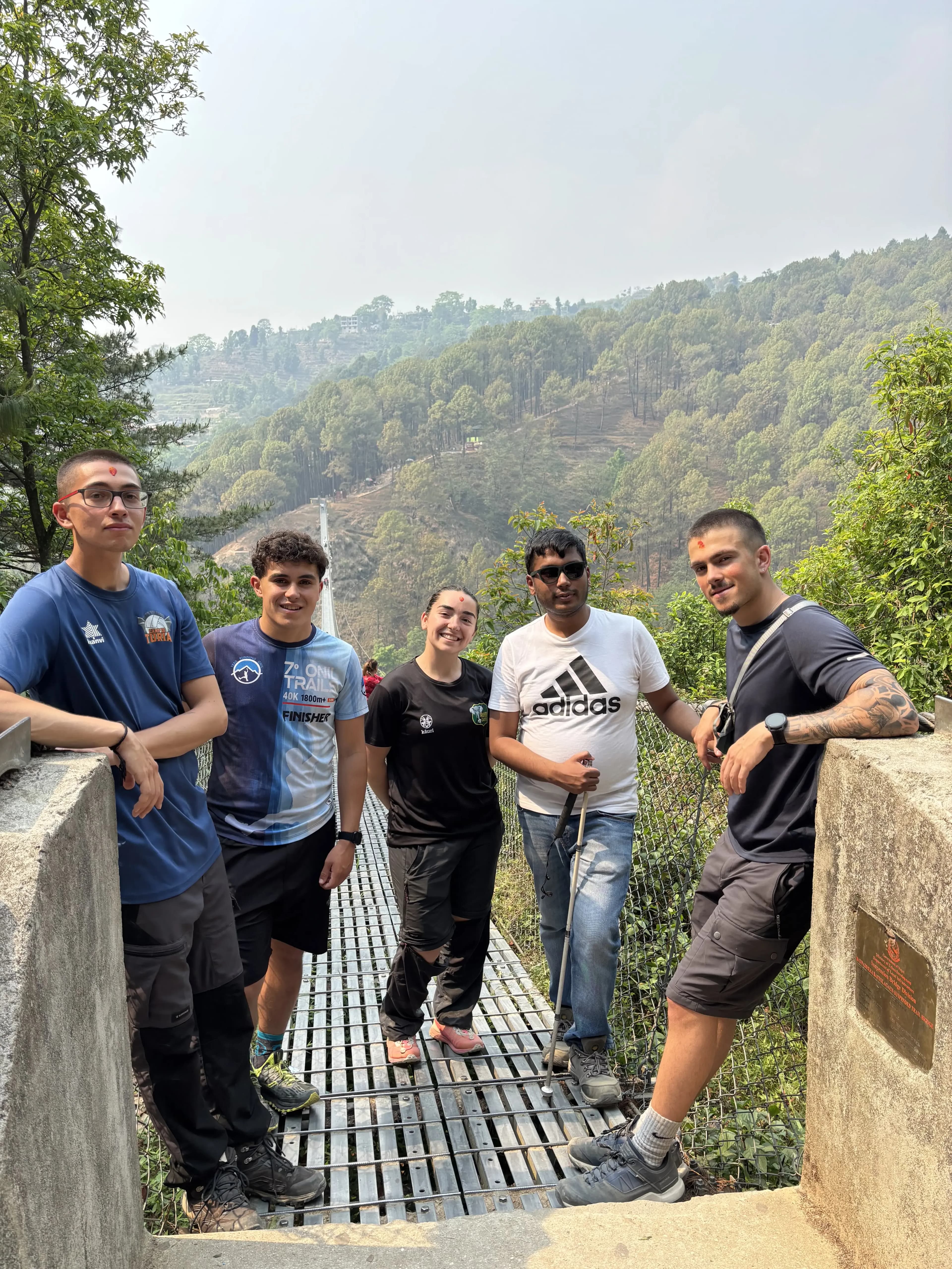 Five hikers pose together at the stone entrance of a narrow suspension bridge, leaning casually on the side walls, with green forested hills stretching into the distance behind them under a hazy sky.