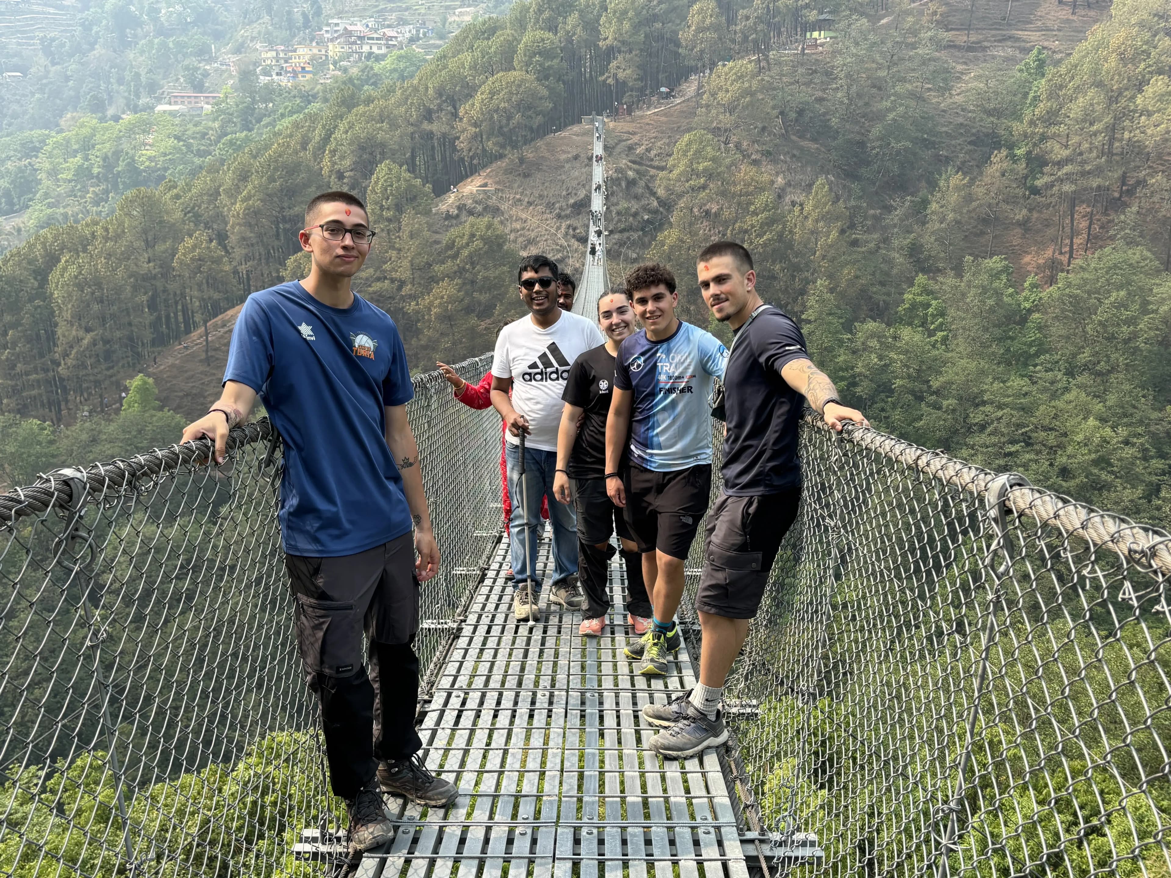 Five hikers stand in a line on a narrow metal suspension bridge high above a green valley, holding the side ropes and smiling, with steep forested hills rising behind them under a hazy sky.