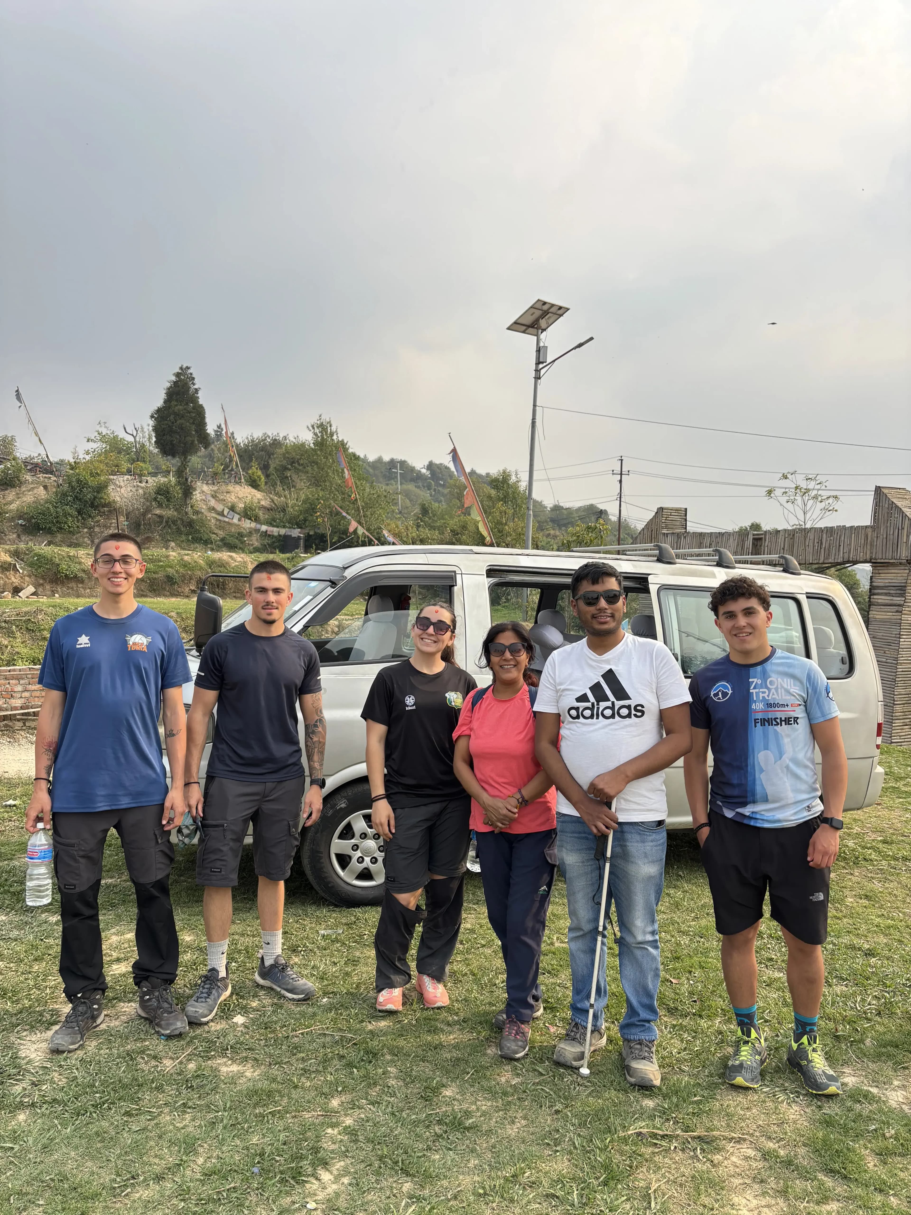 Six hikers stand side by side on a grassy field in front of a white van at the end of a trip, smiling at the camera under a hazy evening sky with low hills and a tall light pole in the background.