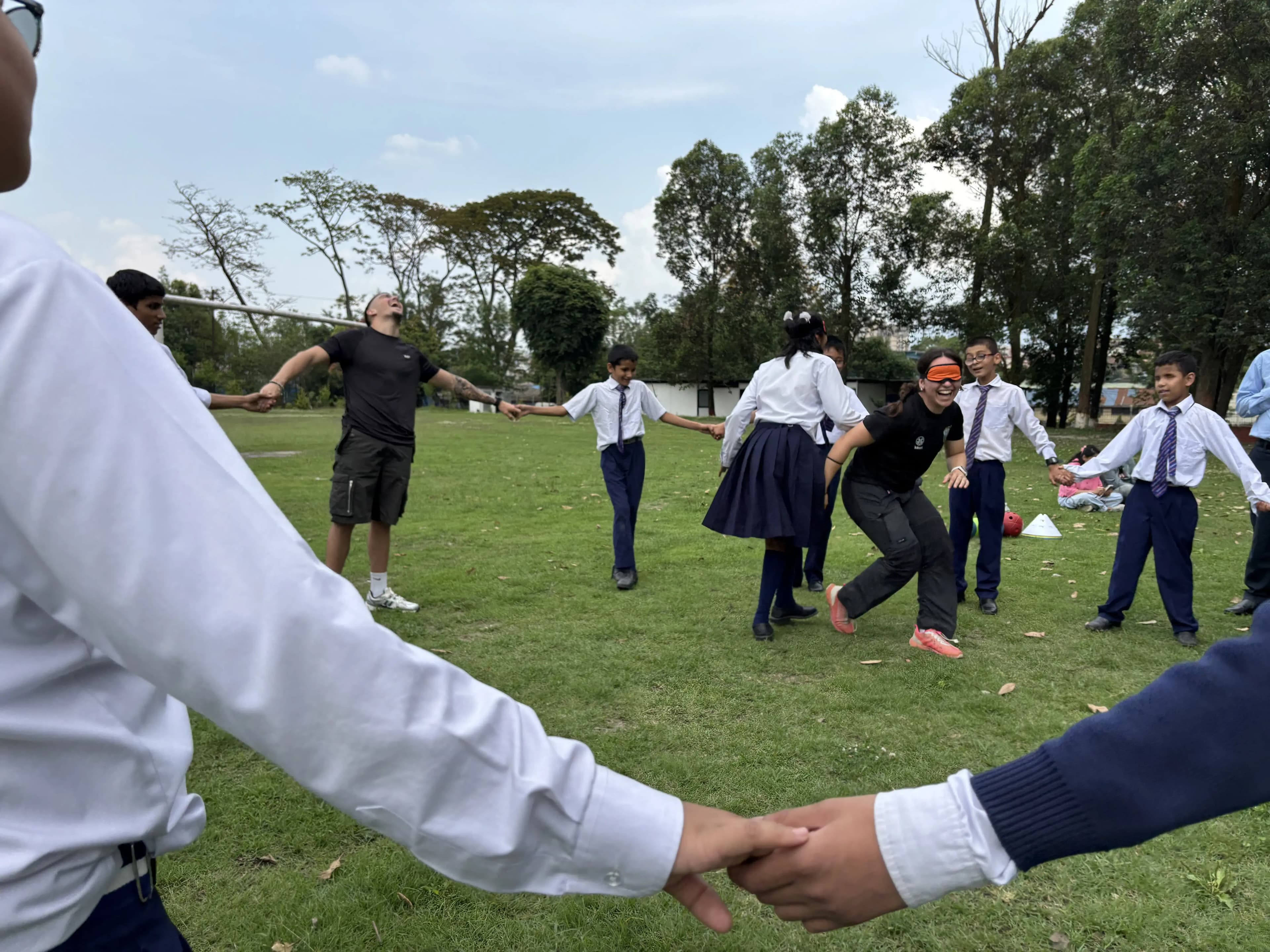 Children in school uniforms and two adults stand hand in hand in a wide circle on a grassy field, playing a game while one adult in the center wears an orange blindfold and moves toward laughing students, with trees and a clear sky in the background.