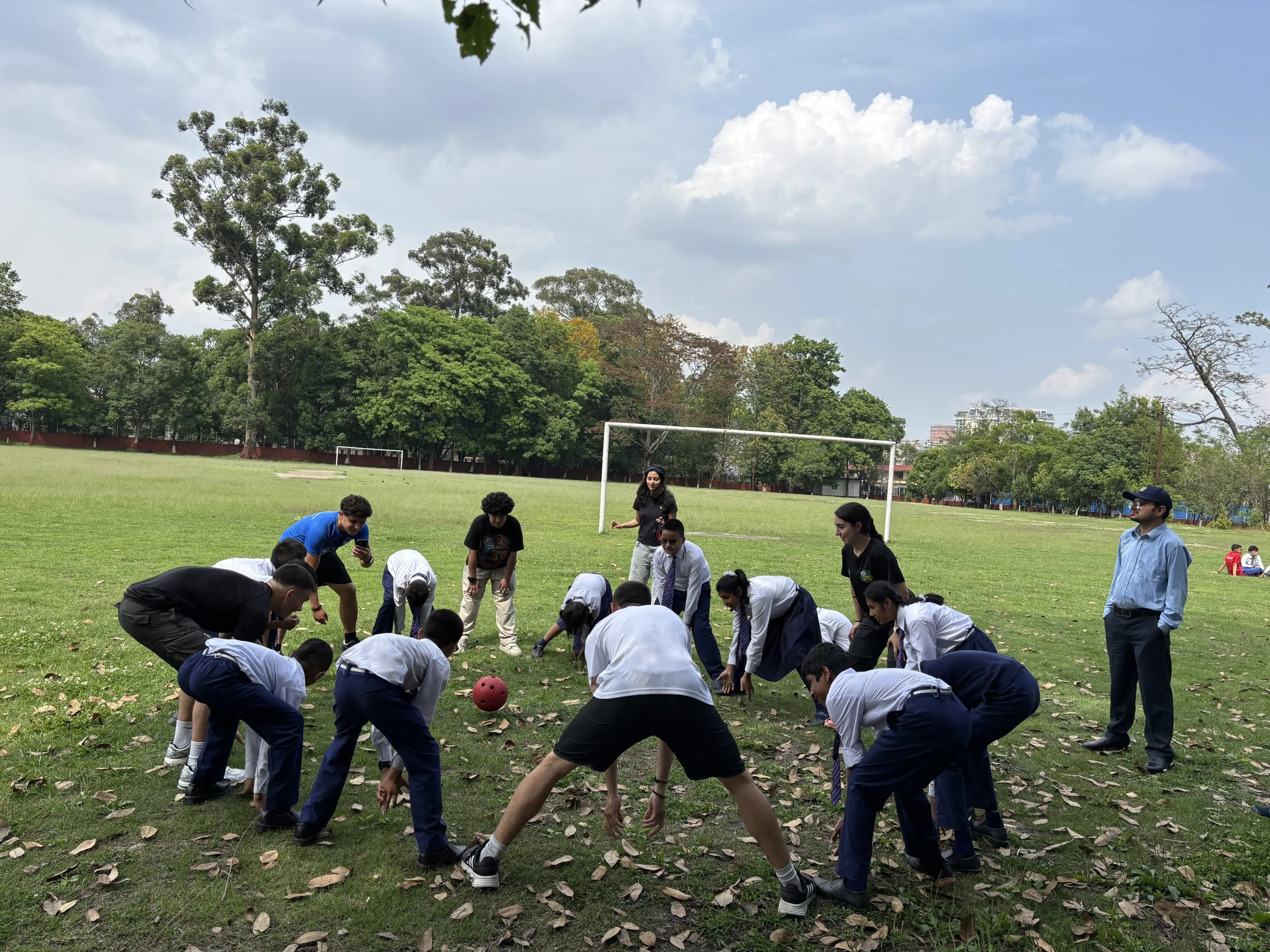 A group of children and an adult stand in a loose circle on a grassy field, all bent over with hands on their knees and heads lowered toward the center, while a bright red ball rests on the ground between them, with tall trees and a blue sky with clouds in the background.