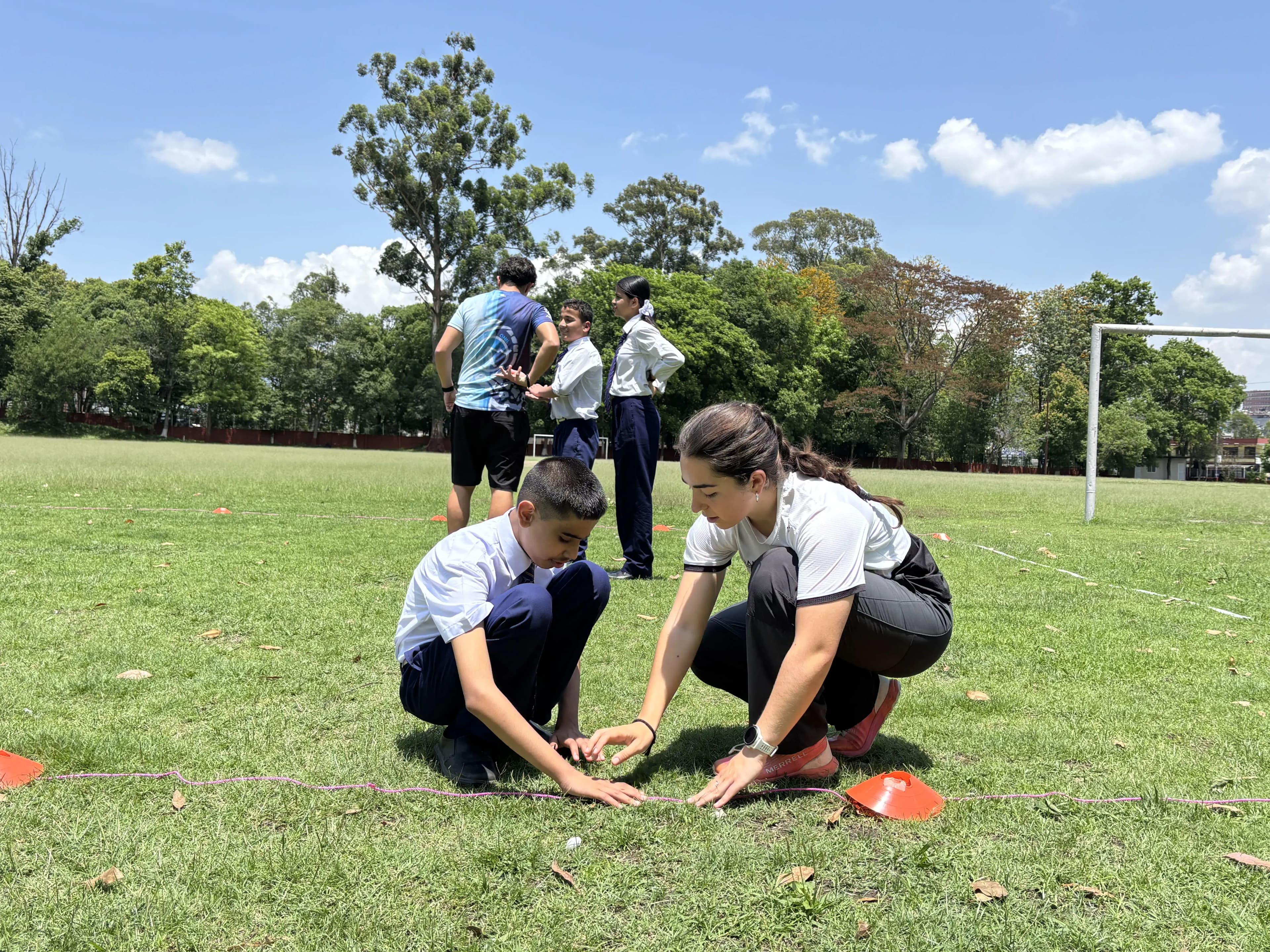 A CFC member and a student kneel on the grass side by side, carefully placing a small ball and feeling the boundary rope between two orange cones as they set up an activity on a sunny field, while other students stand and talk in the background near a goalpost and trees.