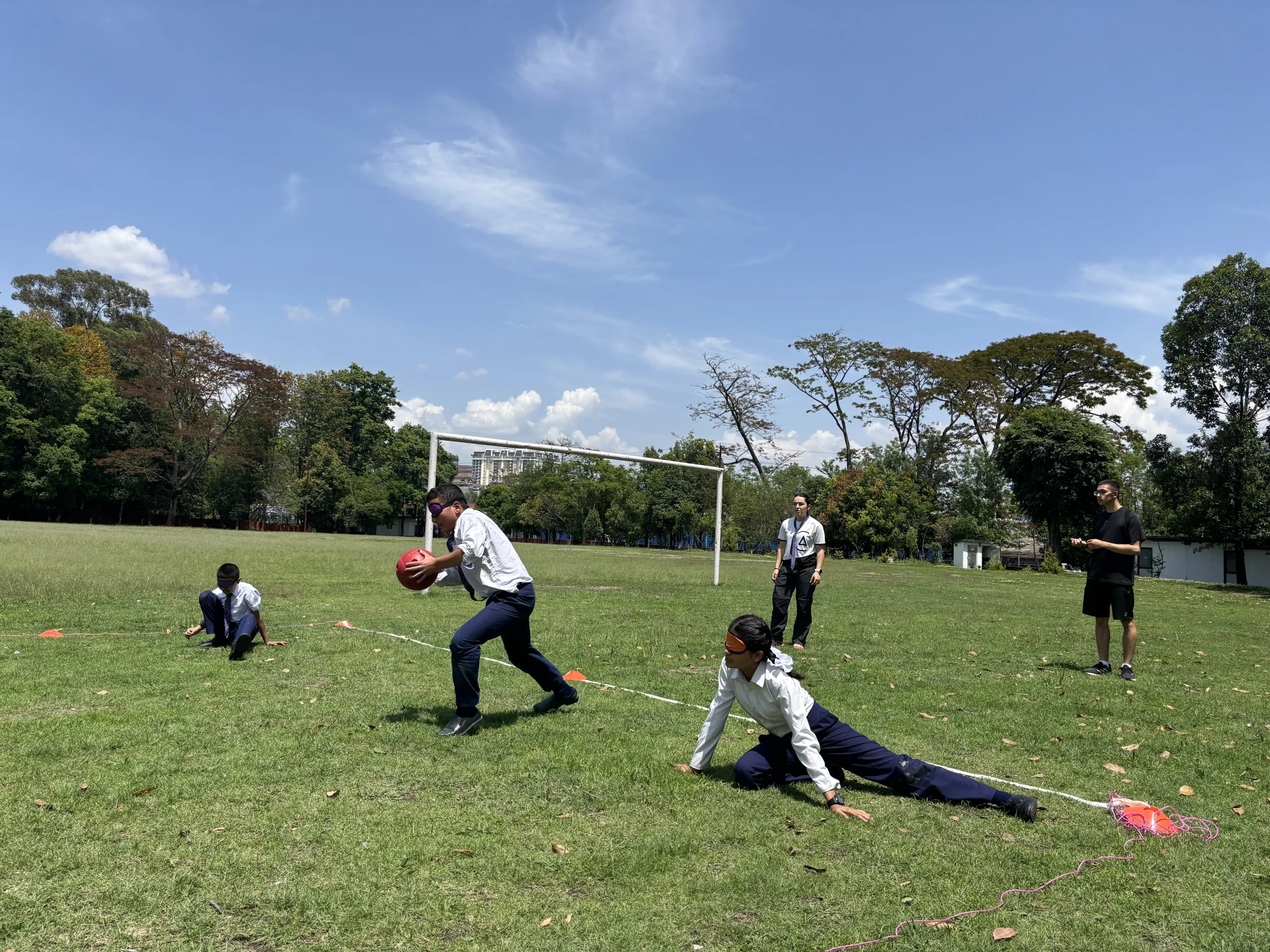A student wearing an eye shade sprints forward on a grassy field holding a large red ball, while another blindfolded student stretches out along a rope on the ground as a defender, and two more students and a CFC member watch from behind under a bright blue sky with scattered clouds.