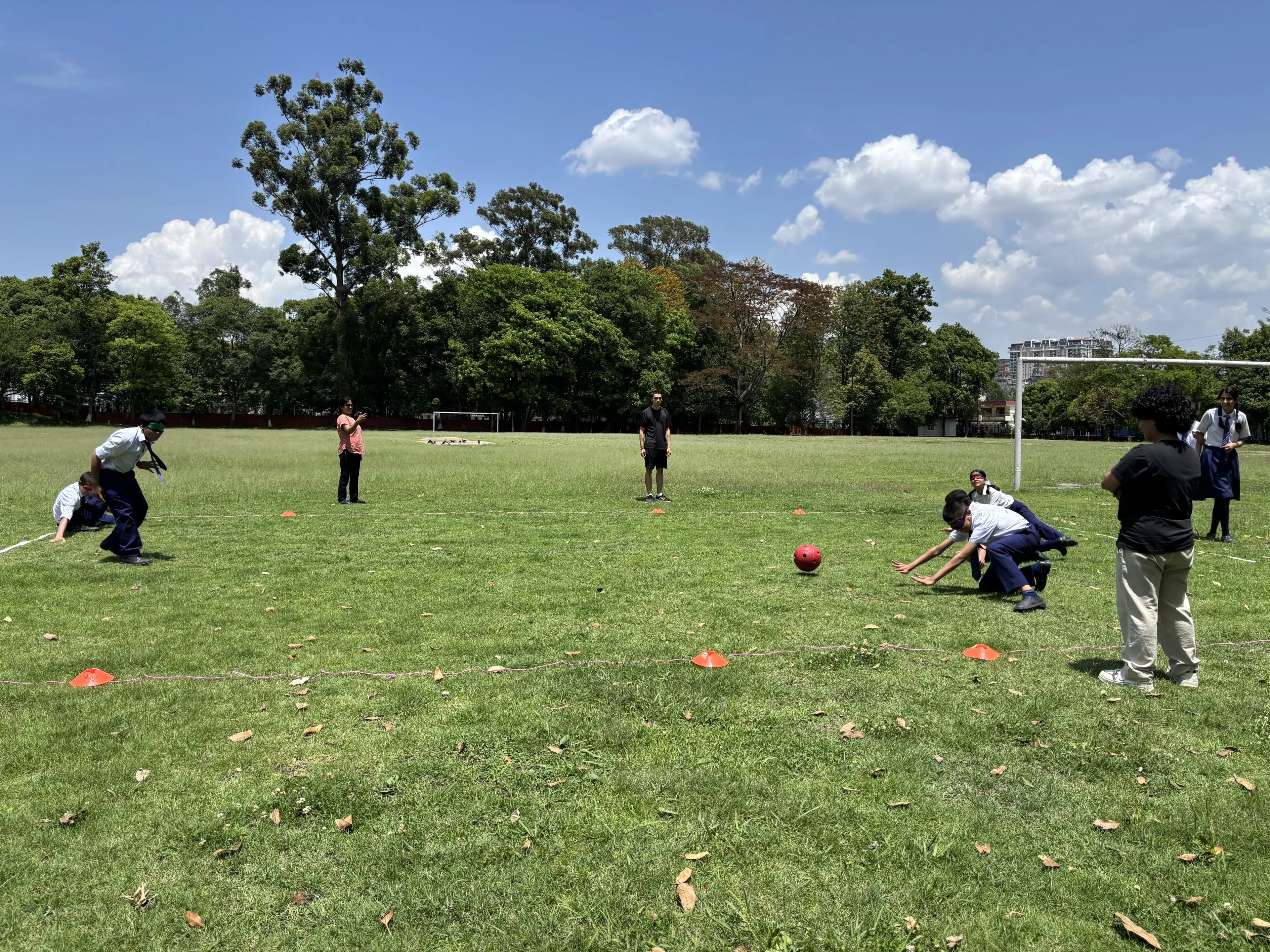 Blindfolded students play goalball on a grassy field, with one student stretched along a rope reaching out as a defender while a red ball rolls toward them, cones marking the court and CFC team and classmates watching under a bright blue sky with scattered clouds.