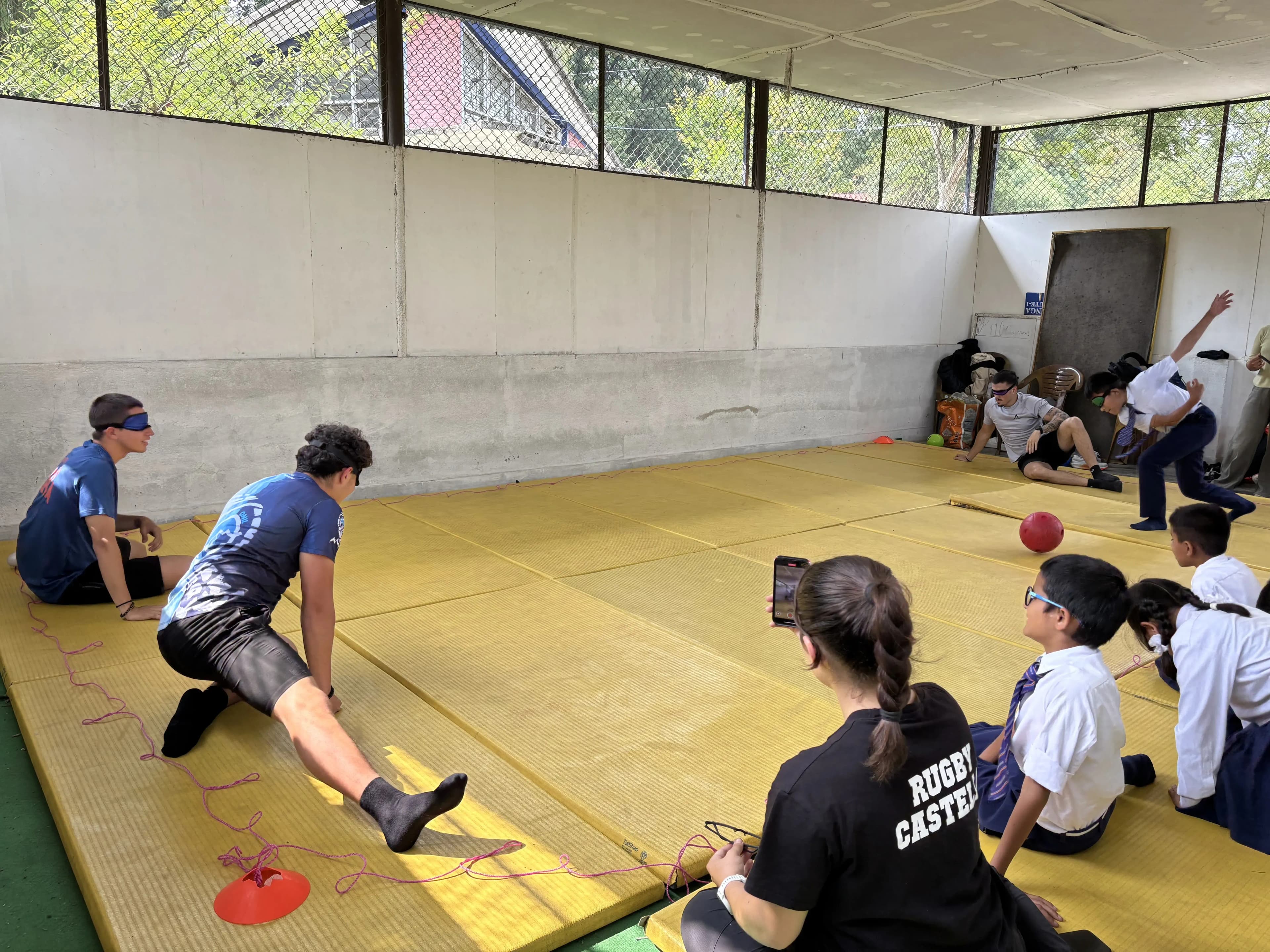 Blindfolded players and schoolchildren sit and kneel around a yellow indoor mat as a coach in sportswear demonstrates goalball technique along a taped boundary line, while a red ball rolls toward a defending student and others watch closely from the sidelines.