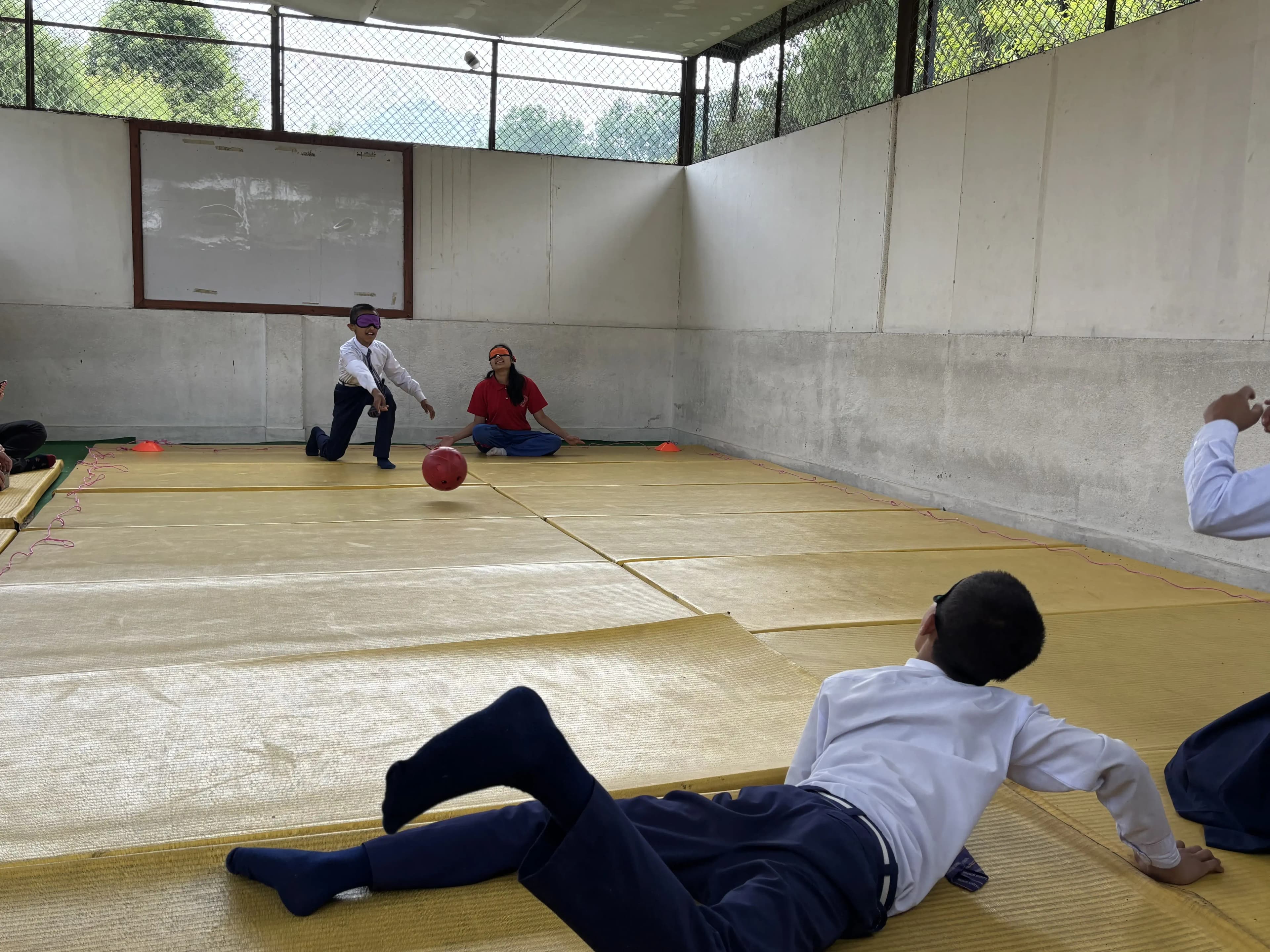 Students in school uniforms practice goalball on yellow mats inside a gym, with one student rolling a red ball toward the opposite end while another lies sideways on the floor as a defender and classmates watch from the sidelines.