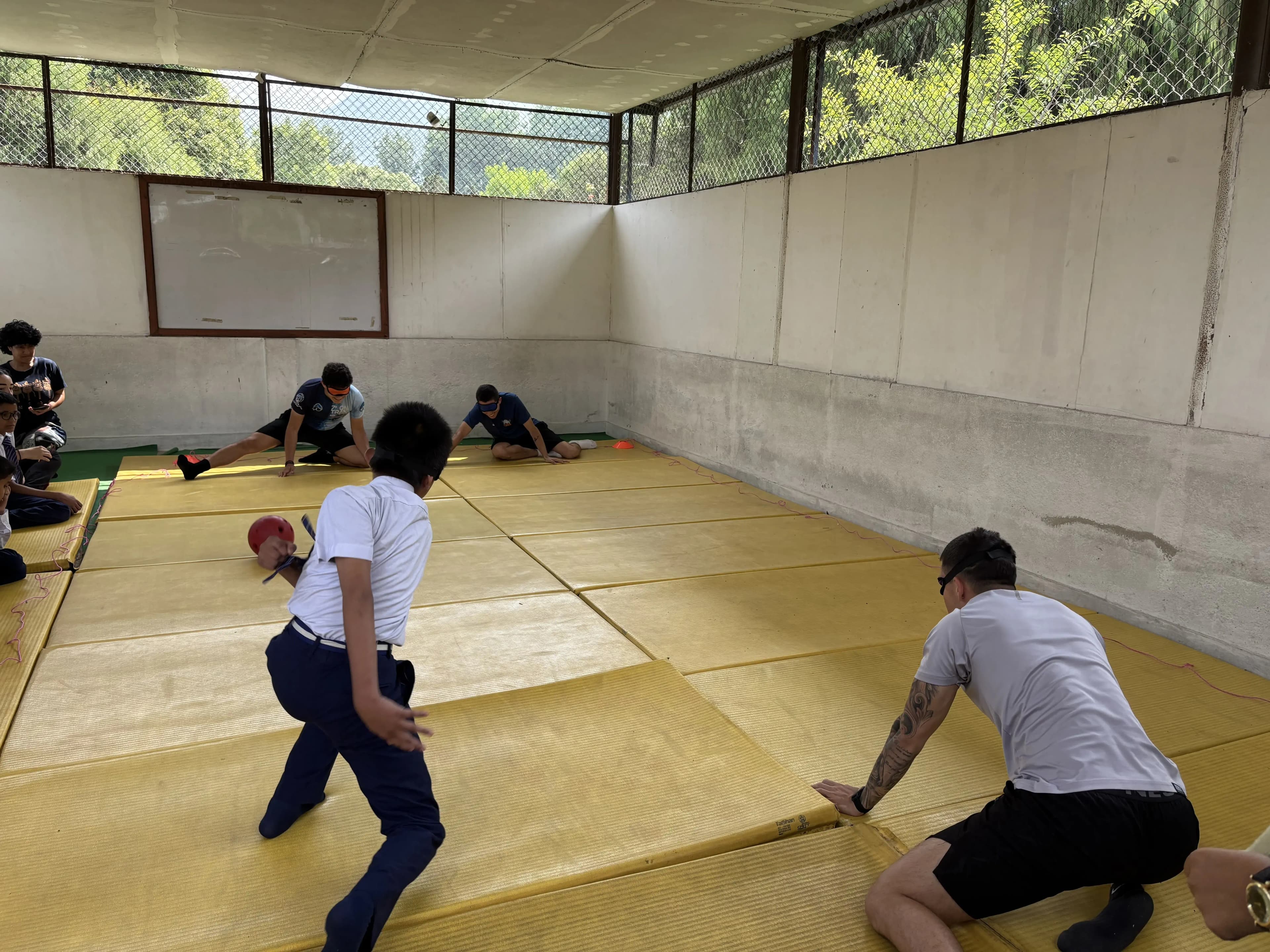 People playing goalball on yellow mats inside a long indoor court, with two players in the foreground moving toward the ball while several others sit or kneel along the sides watching.