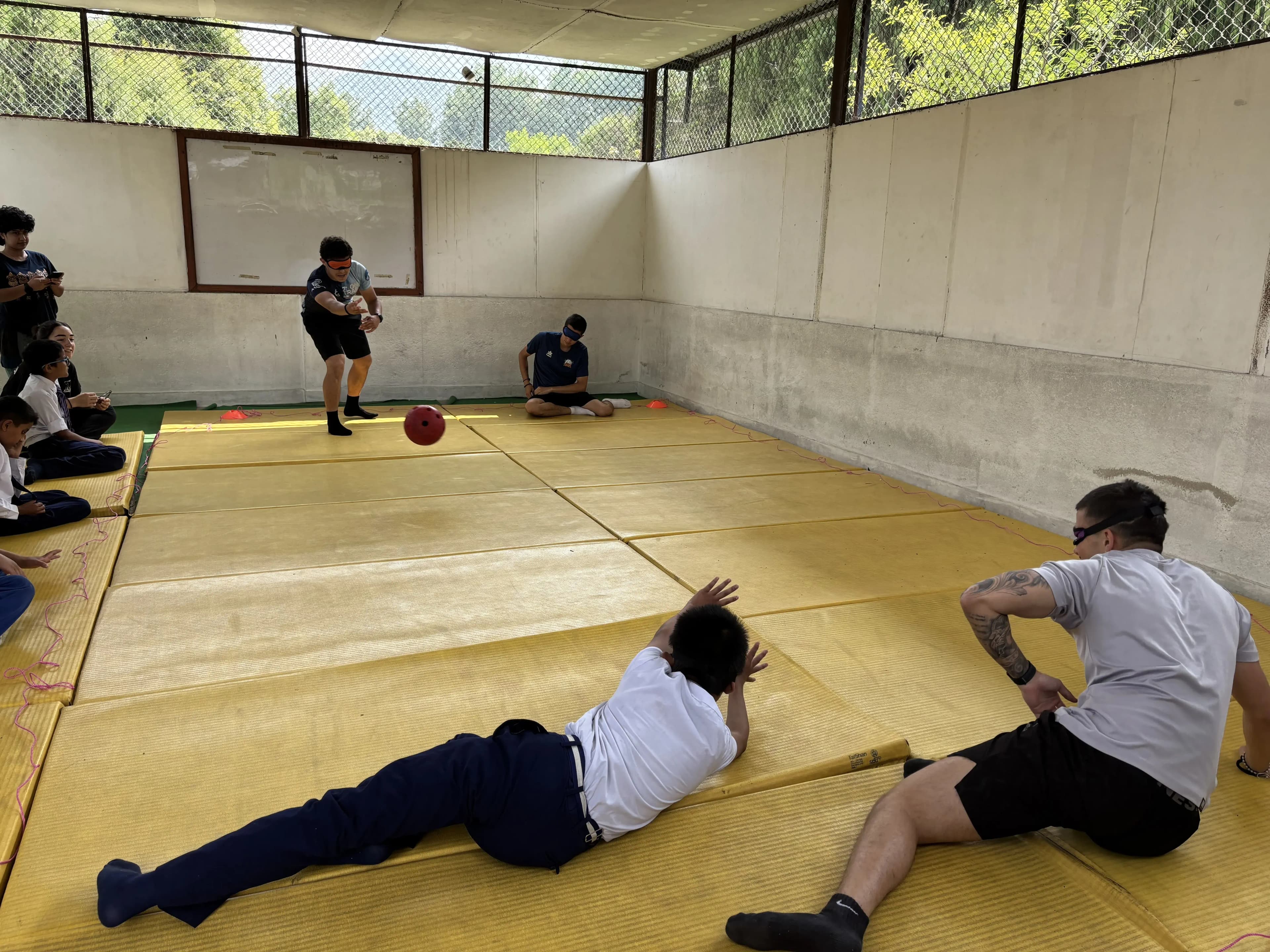 Goalball game on yellow mats in a long indoor court, with one player in the foreground stretched out on the floor blocking, another player sitting nearby ready to move, and a third player at the far end preparing to throw the ball while teammates and CFC members sit along the side watching.