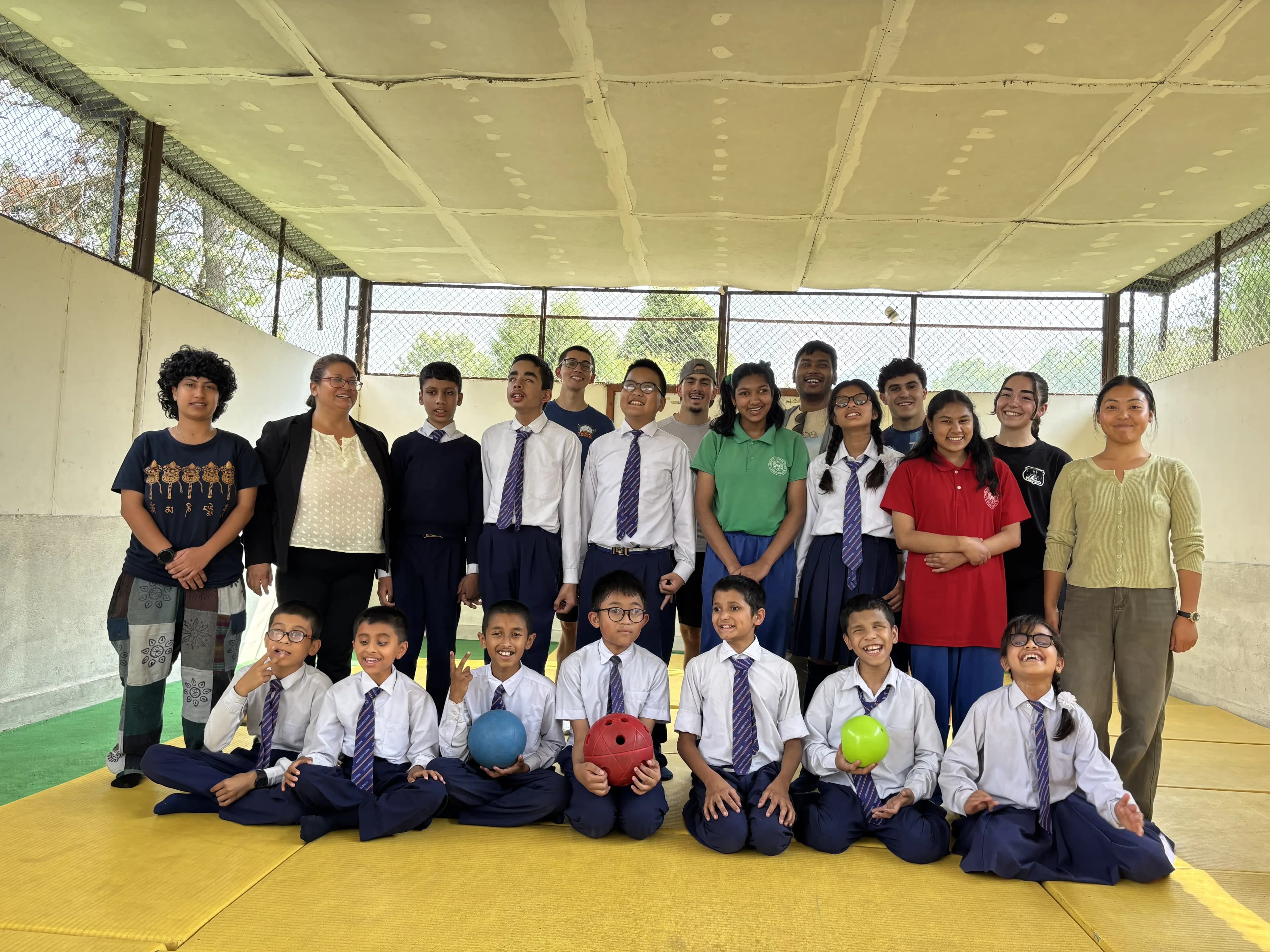 Group photo of goalball players and coaches in an indoor court, with younger students in school uniforms holding goalballs seated in the front row and older students and CFC members standing behind them, all smiling on the yellow mat.