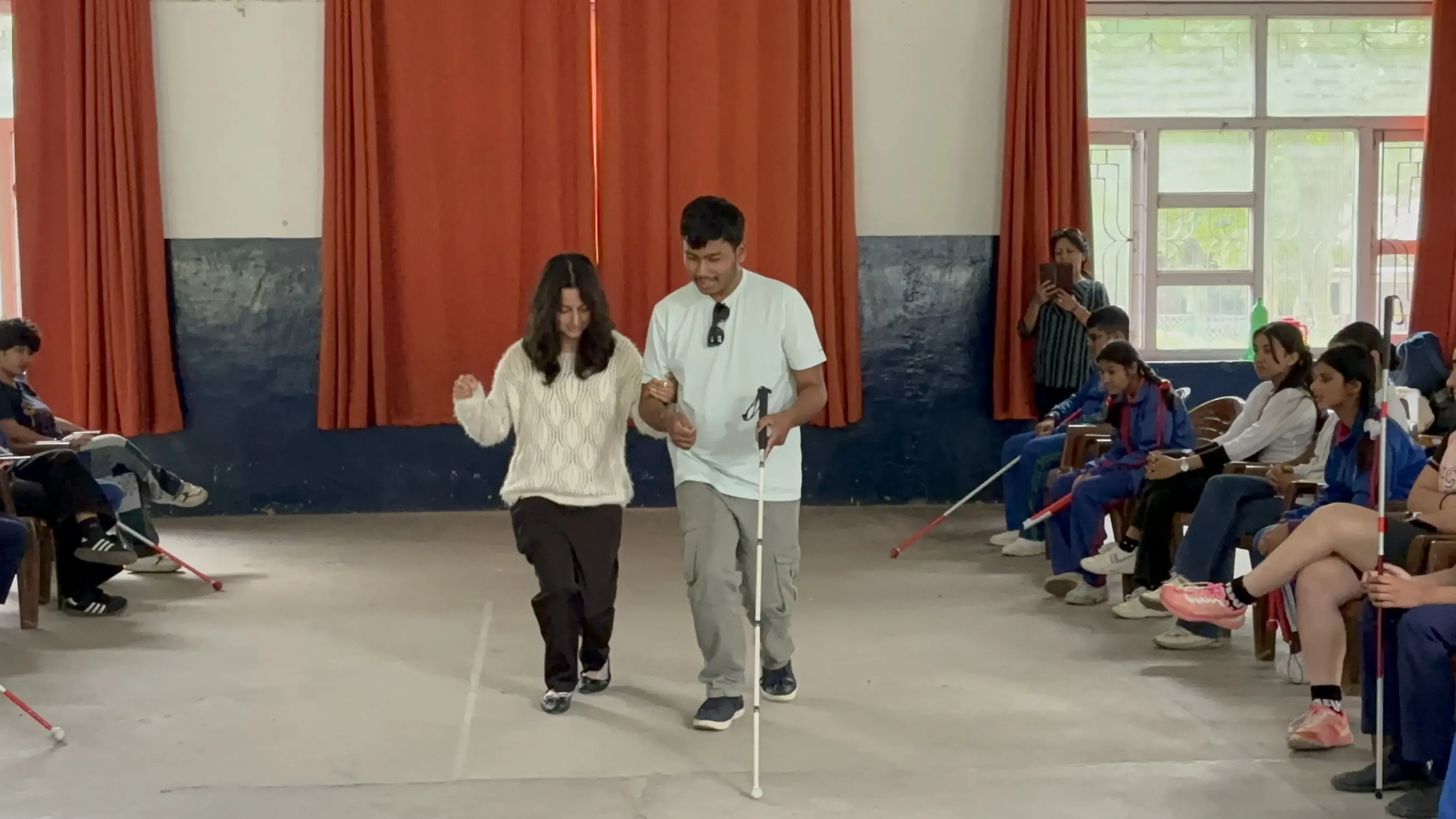 Indoor mobility skills training session where a blindfolded female practices walking with guide support and a white cane in the center of the room, while a group of participants with white canes sit in chairs along both sides watching.