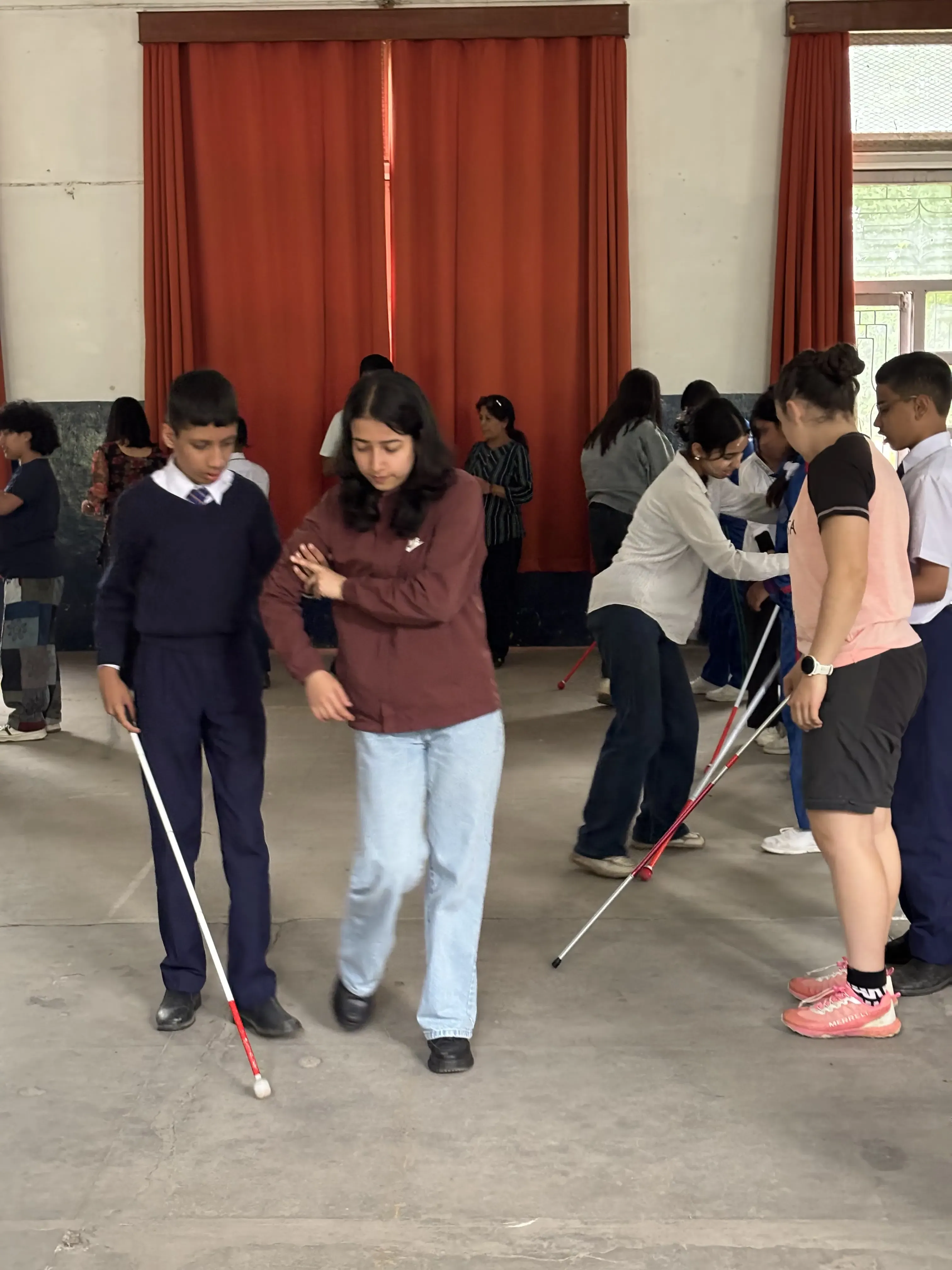 Indoor orientation and mobility training session where a female guides a student using a white cane across the room, while other pairs of participants nearby also practice walking with white canes.