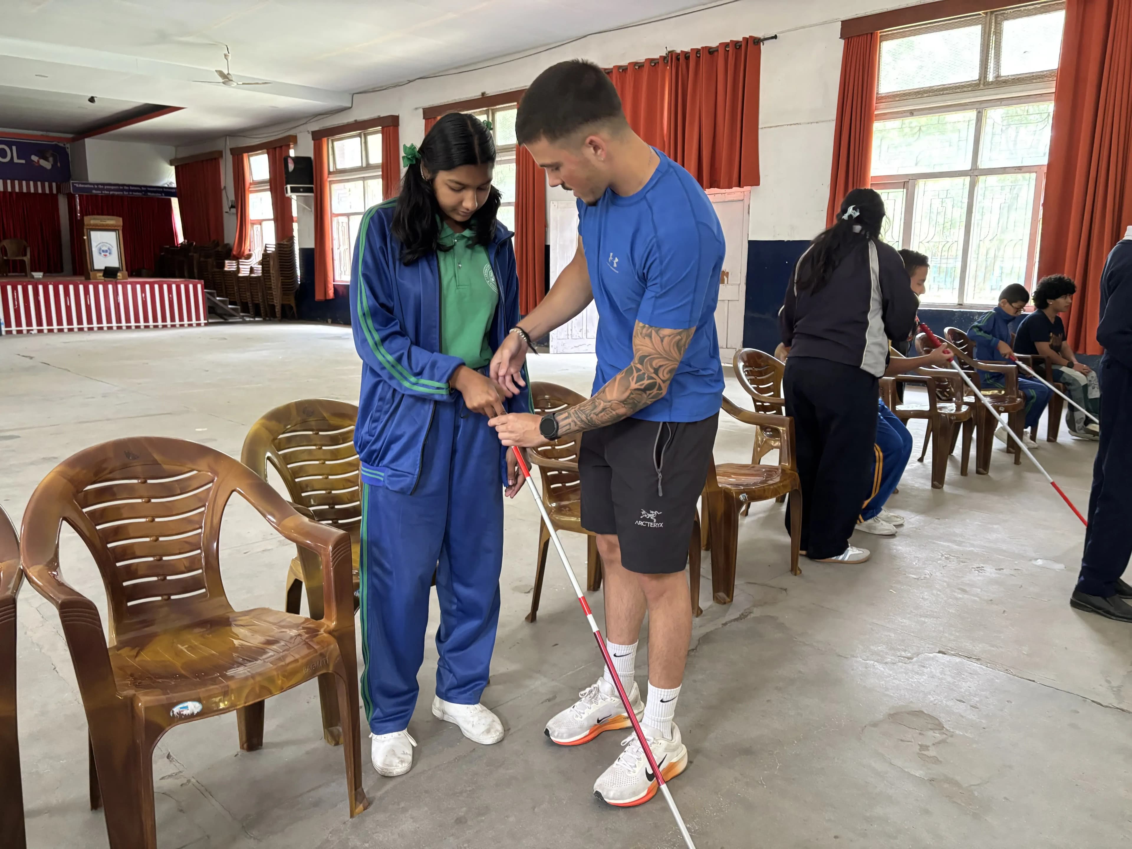 CFC member demonstrating proper white cane grip and guiding technique to a student in a tracksuit during an indoor orientation and mobility training session, with other participants practicing with canes in the background.