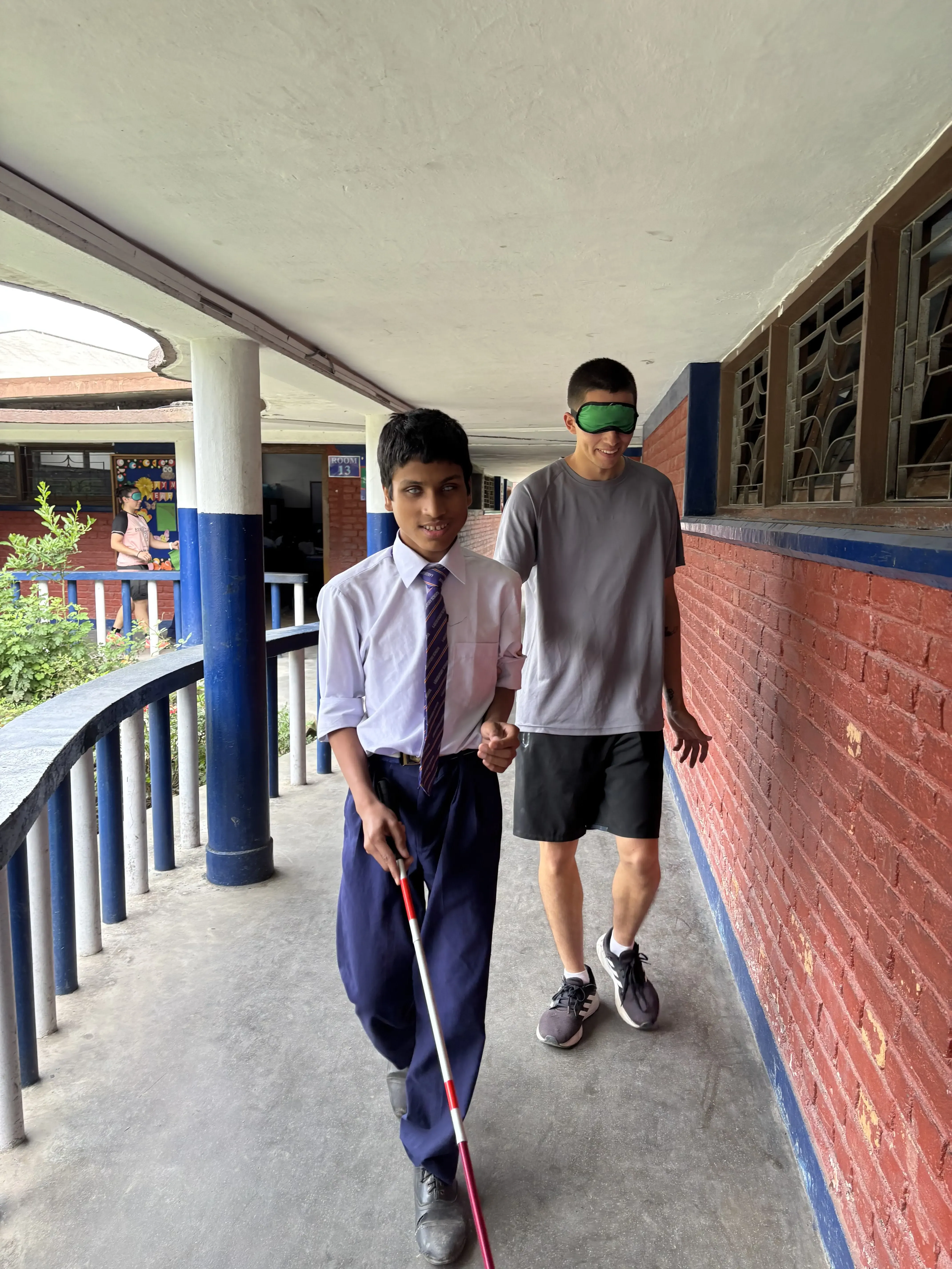 Student with a visual disability in school uniform guiding a blindfolded pair guide along an outdoor corridor, using a white cane and holding the person's arm as they walk beside a brick wall and railing.
