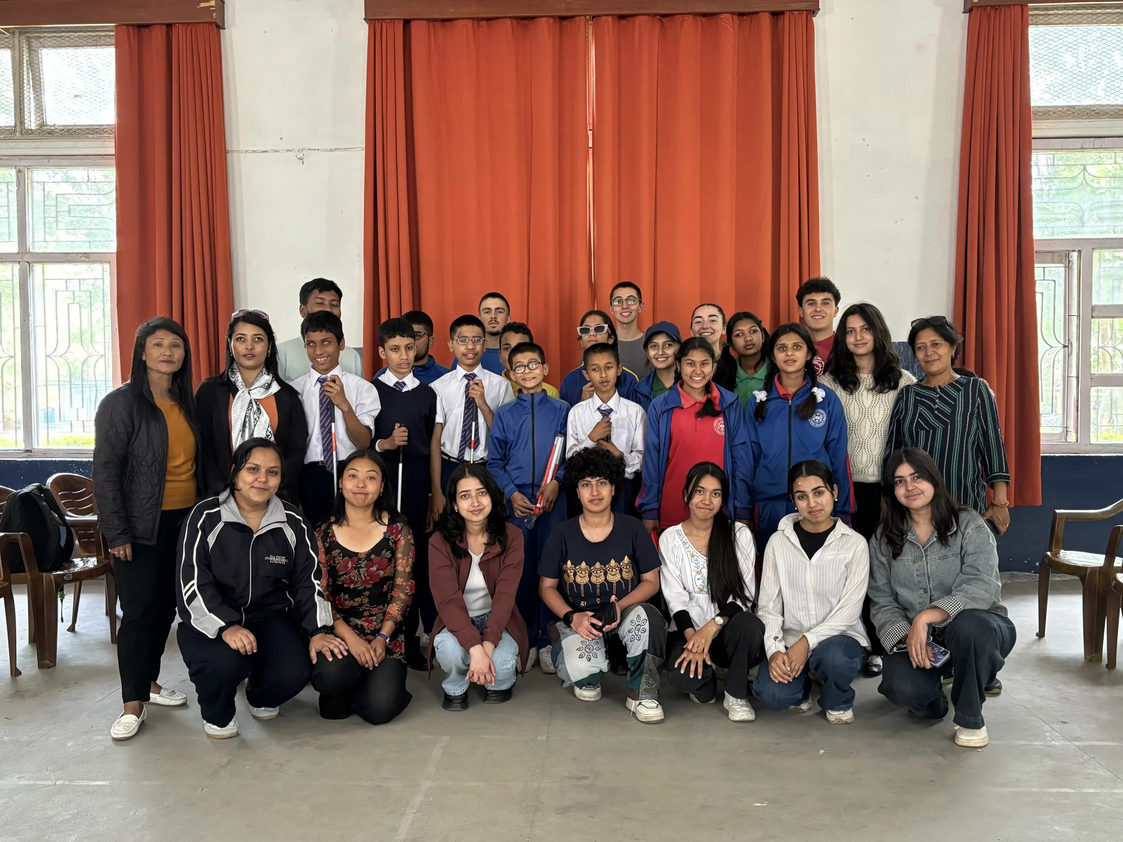 Group photo of students and CFC members from the guiding techniques and mobility skills workshop, with younger students in school uniforms standing in the middle and CFC members in casual clothes kneeling and standing around them in a hall with large orange curtains.