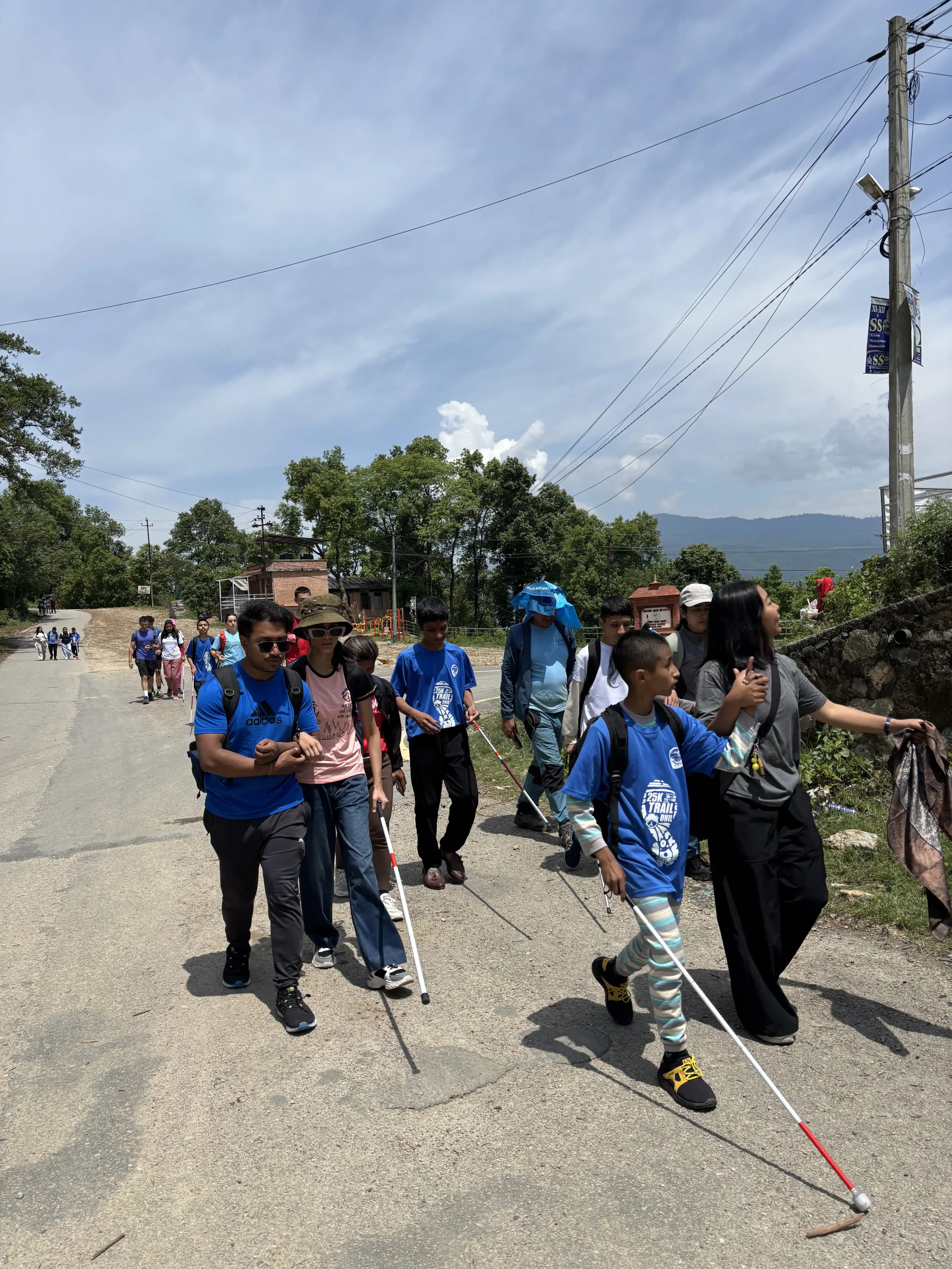 Inclusive Hiking day where students using white canes practice road-crossing and outdoor orientation skills along a sunny village street, each paired with a sighted guide as they walk in a line past houses, trees, and utility poles.