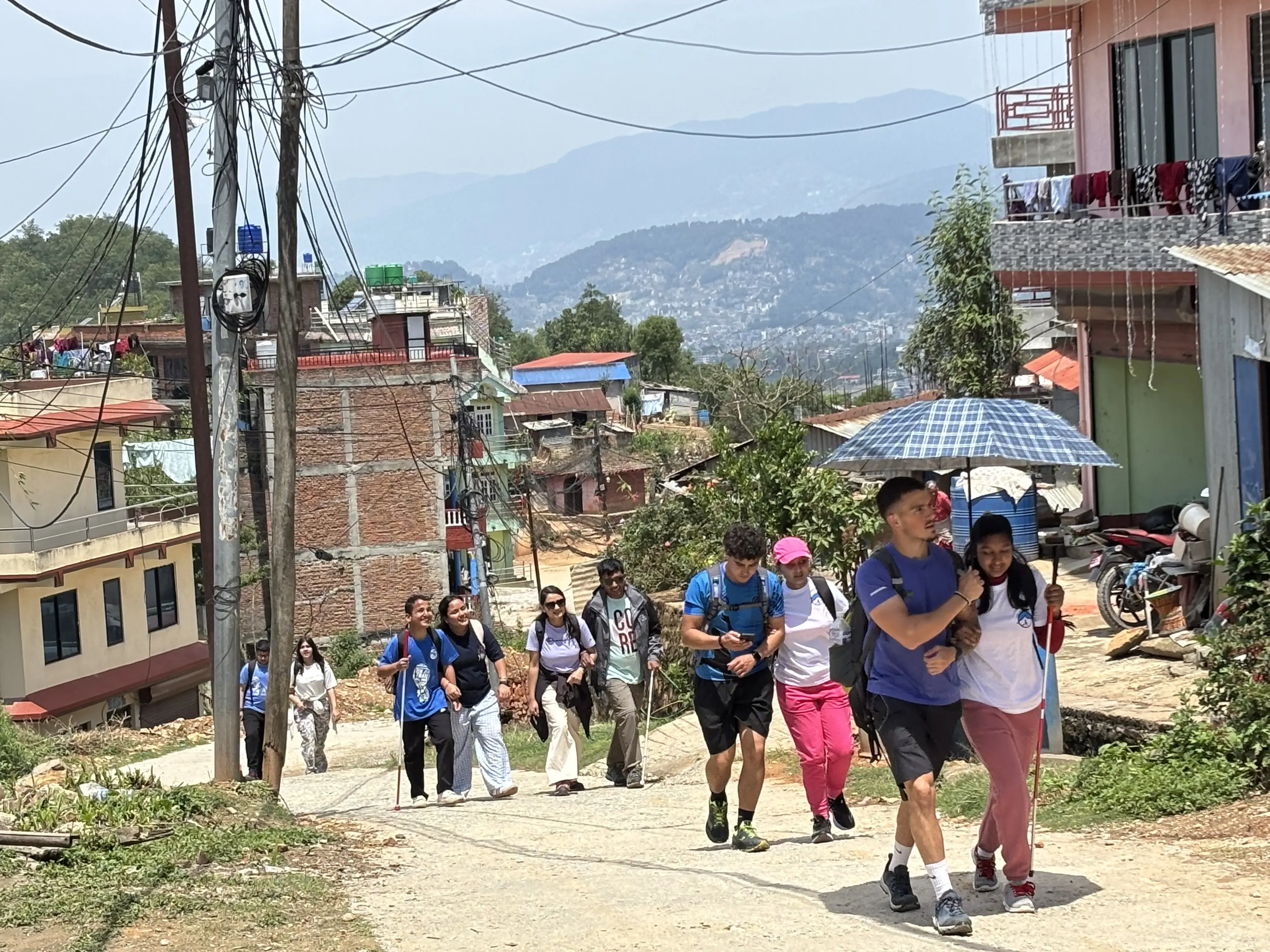 Inclusive Hiking day where participants practicing outdoor guiding techniques as pairs walk up a sunny hillside lane in a village, with one person leading under an umbrella while others follow behind between houses, power poles, and distant mountains.