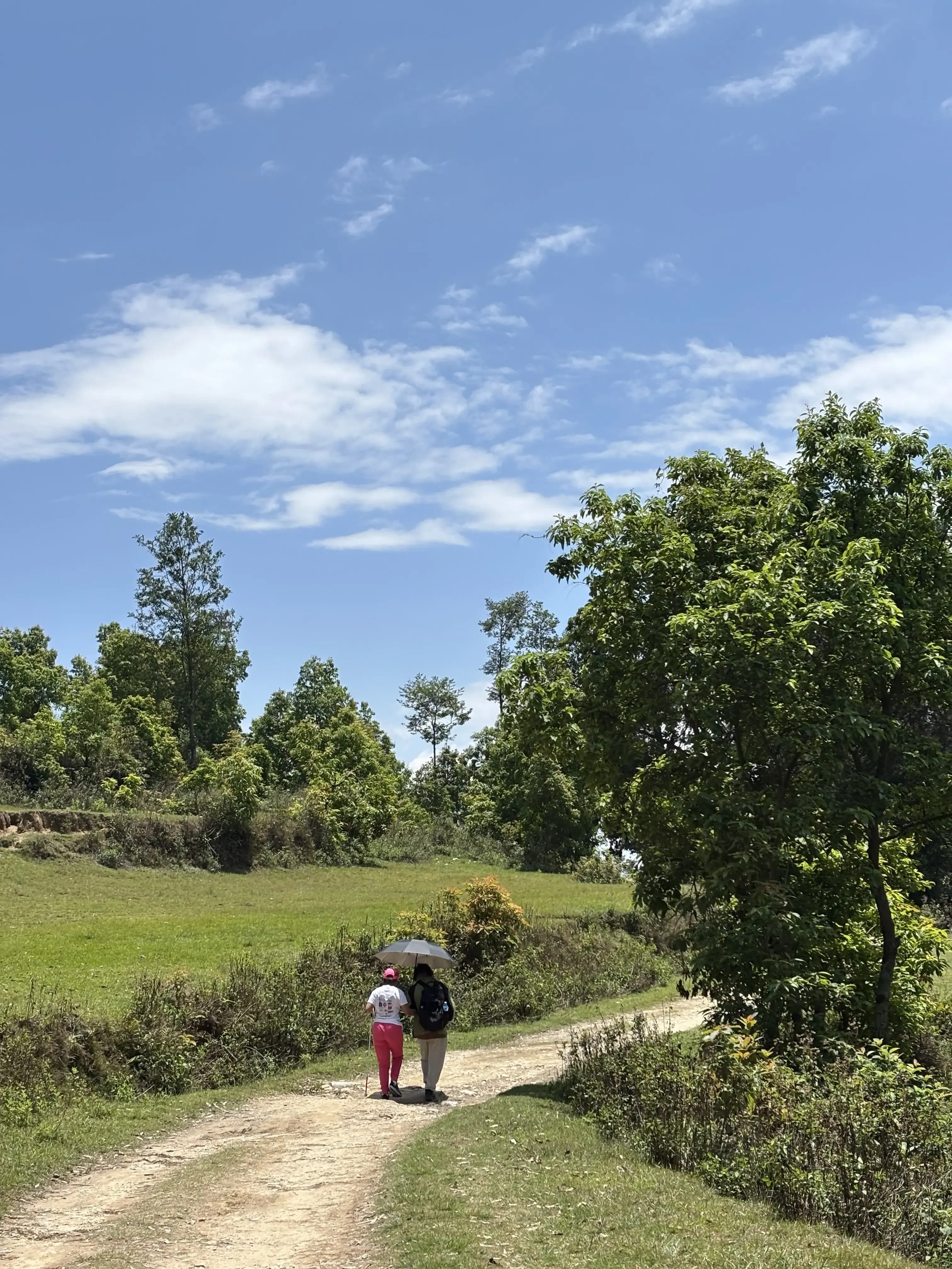 Inclusive Hiking day where two participants walk together under an umbrella along a winding dirt path through green fields and trees on a bright, clear day, practicing outdoor guiding skills in a rural setting.