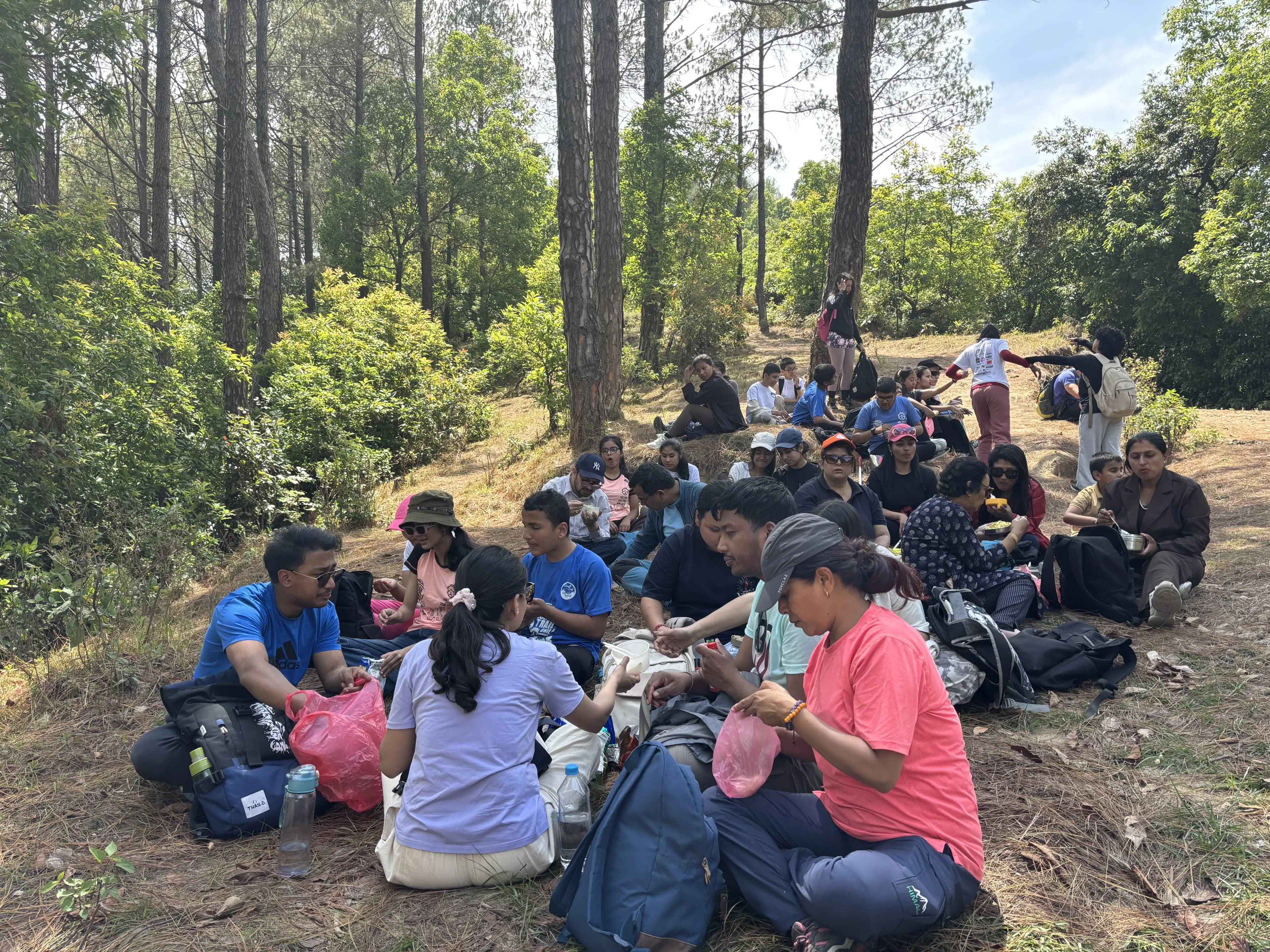Inclusive Hiking day where a group of participants resting together on a forest hillside during an outdoor excursion, sitting on the ground among trees and backpacks while sharing snacks and talking.