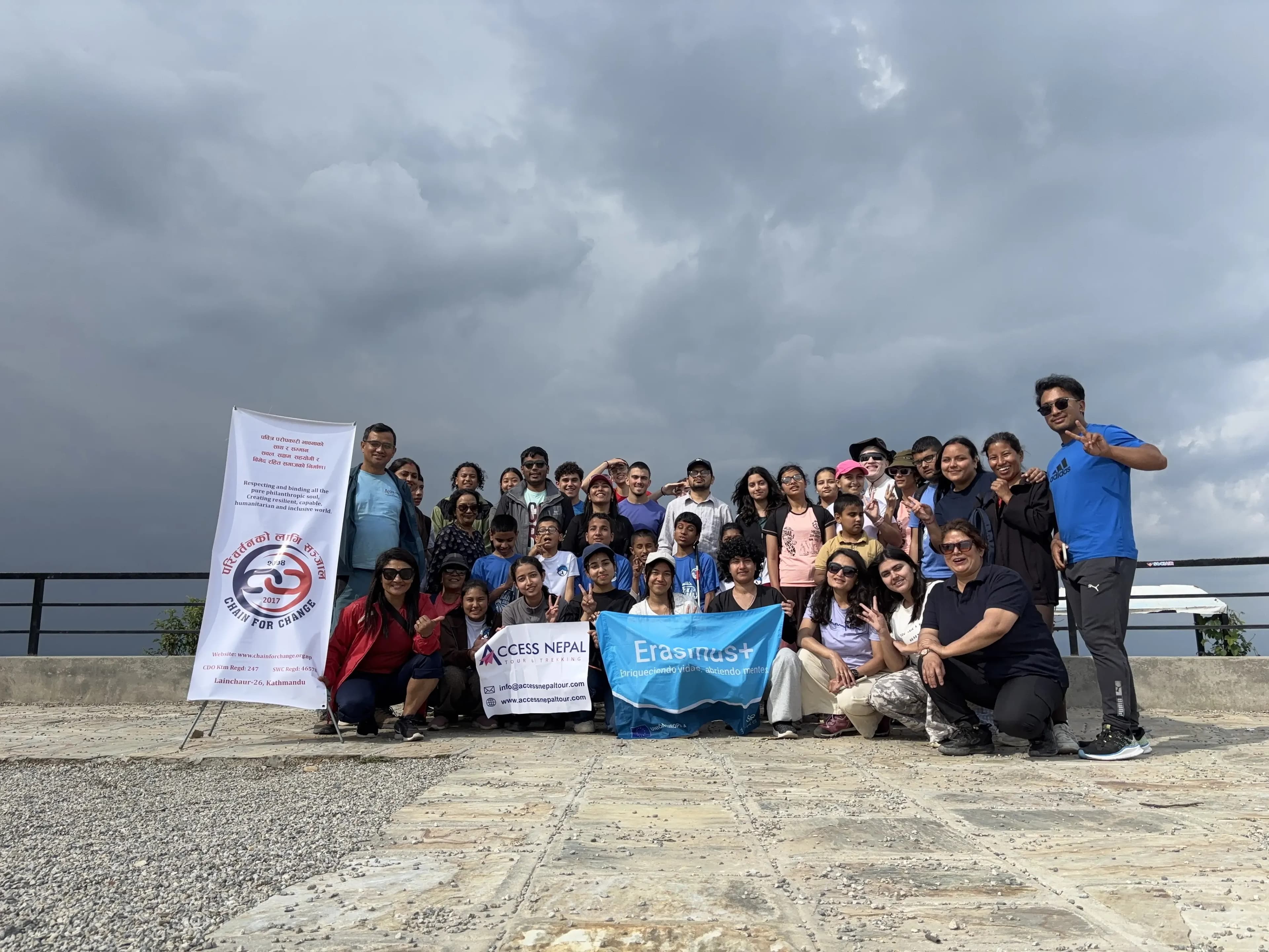 Group photo of participants holding Access Nepal and Erasmus+ banners on an open terrace under a cloudy sky, with people of different ages smiling and posing together in front of a metal railing.