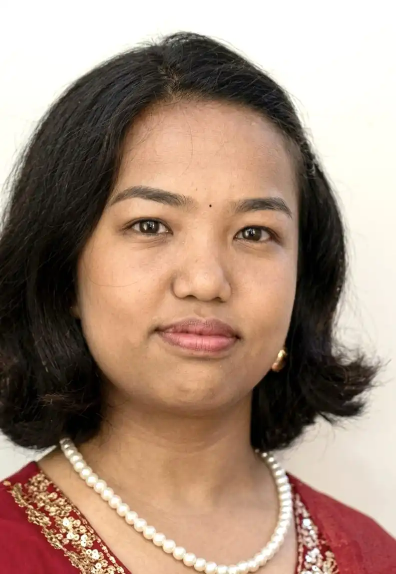 Woman with medium-brown skin and shoulder-length black hair, wearing a pearl necklace, gold earrings, and a red outfit with gold embroidery, facing the camera with a calm expression against a plain light background.