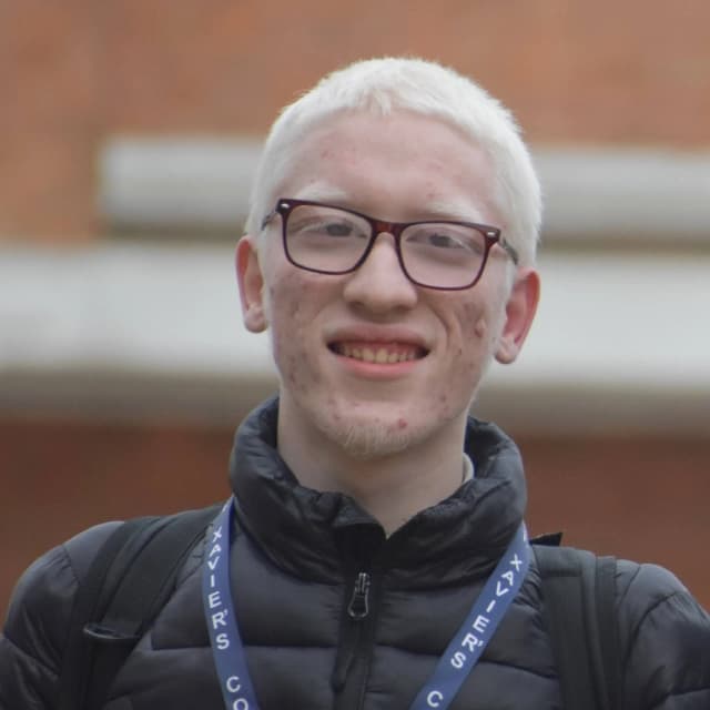 Young man with very light skin and short white hair, wearing dark rectangular glasses and a black puffer jacket with backpack straps and a blue lanyard around his neck, smiling slightly with an outdoor background.