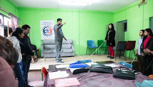 In this image, the Founder & director of CFC Mr. Abhishek Shahi and Co-founder and Treasurer Ms. Sanjiya Shrestha are showing and explaining fellow trainees how to use their white canes correctly on the stage. Trainees with visual disability are being guided by their pairs in learning, understanding and following the steps being demonstrated to them. There is a sofa and chair placed above the stage and a banner of Chain For Change is also hanged on the wall.