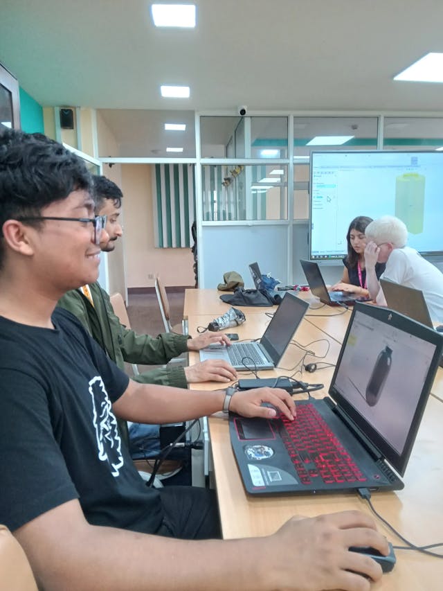A picture where one of the dedicated team has completed designing a bottle in his laptop, on the screen a bottle is observed and in the background intern design instructor is interacting with other members of the class.