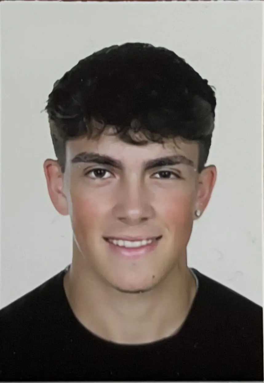 Young man with light-tan skin and short dark curly hair, wearing small earrings and a black top, smiling slightly at the camera against a plain light background.