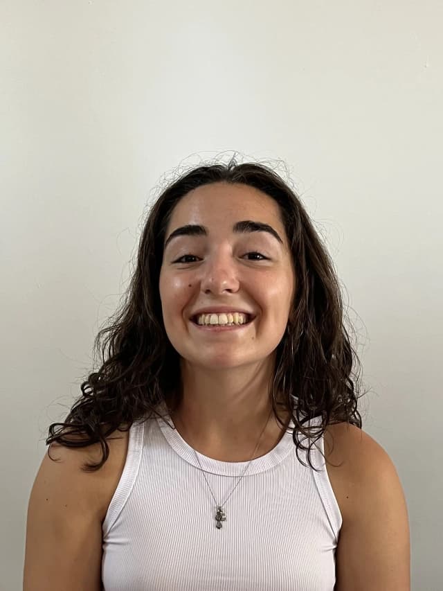 Young woman with light-tan skin and shoulder-length wavy dark hair, smiling broadly and showing her teeth, wearing a white sleeveless top and a small pendant necklace, in front of a plain light-colored wall.