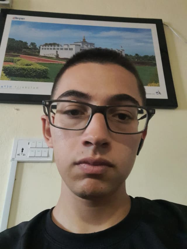 Young man with light-brown skin, very short dark hair, and rectangular black glasses, wearing a black shirt, facing the camera with a neutral expression; a framed landscape picture and a light switch are on the wall behind him.