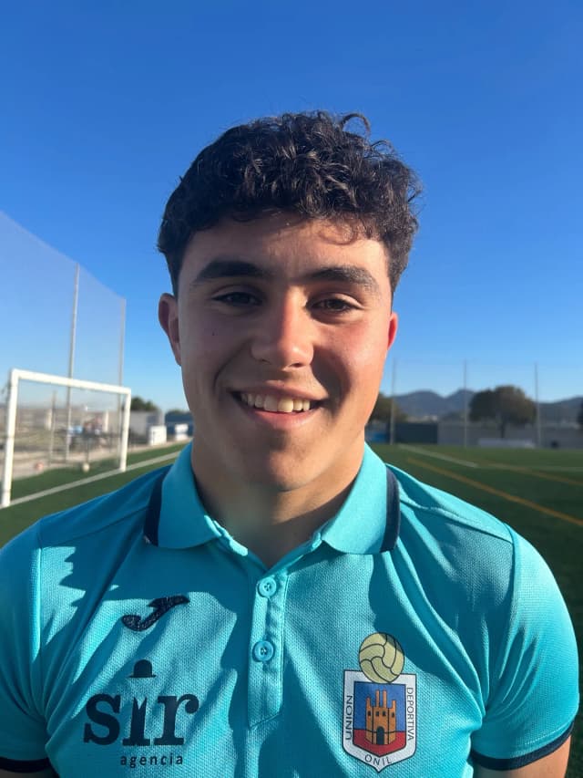 Young man with light-tan skin and short curly dark hair, smiling and wearing a turquoise soccer polo shirt a crest featuring a volleyball and a tower, standing on an outdoor soccer field under a clear blue sky.